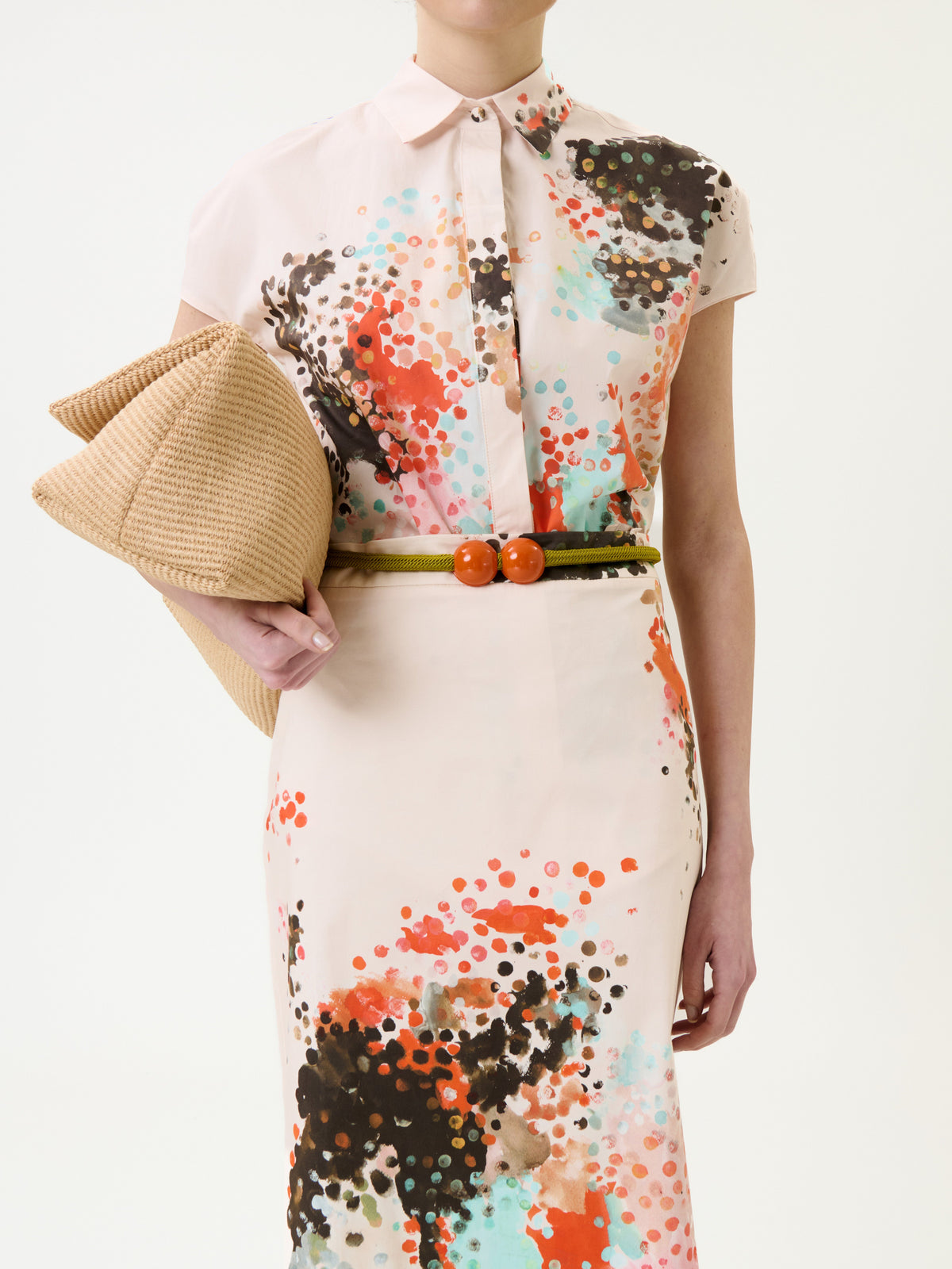 Woman wearing the Melia Skirt, featuring a colorful paint splatter pattern, styled with a green belt with orange accents, and holding a large woven beige clutch. Only her torso and part of her arms are visible.