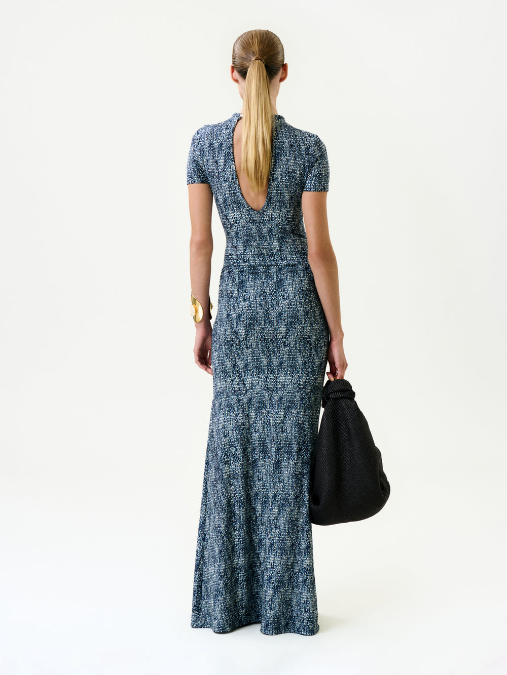 A woman with long hair in a low ponytail wears the Yara Dress, a short-sleeved blue patterned maxi from Colombia with a low scoop back. She faces away, holding a large textured black bag against a plain white background.