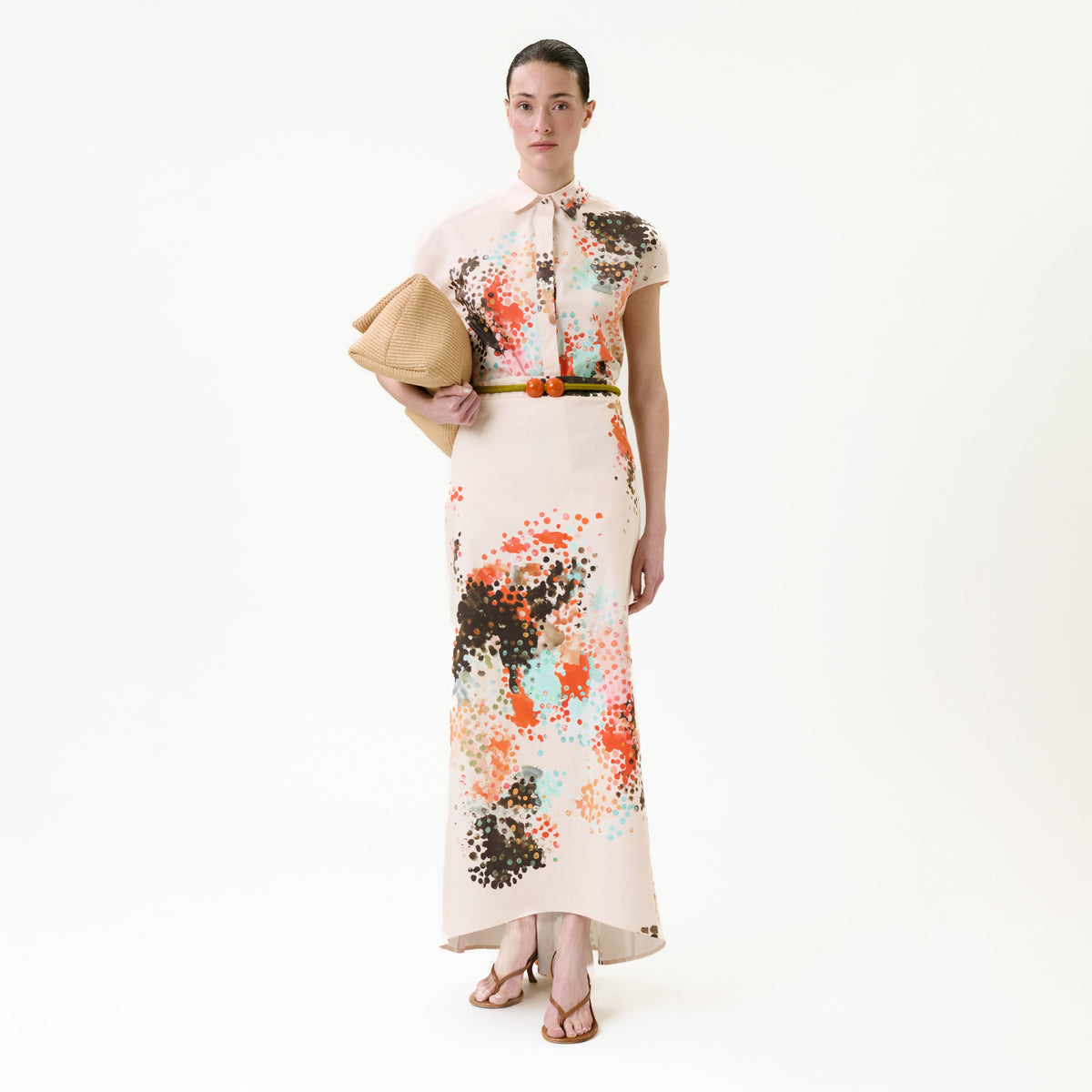 A woman wearing the Melia Skirt, a long, light-colored skirt with abstract multicolored spots, styled with a brown belt and sandals, holding a tan hat and standing against a plain white background.