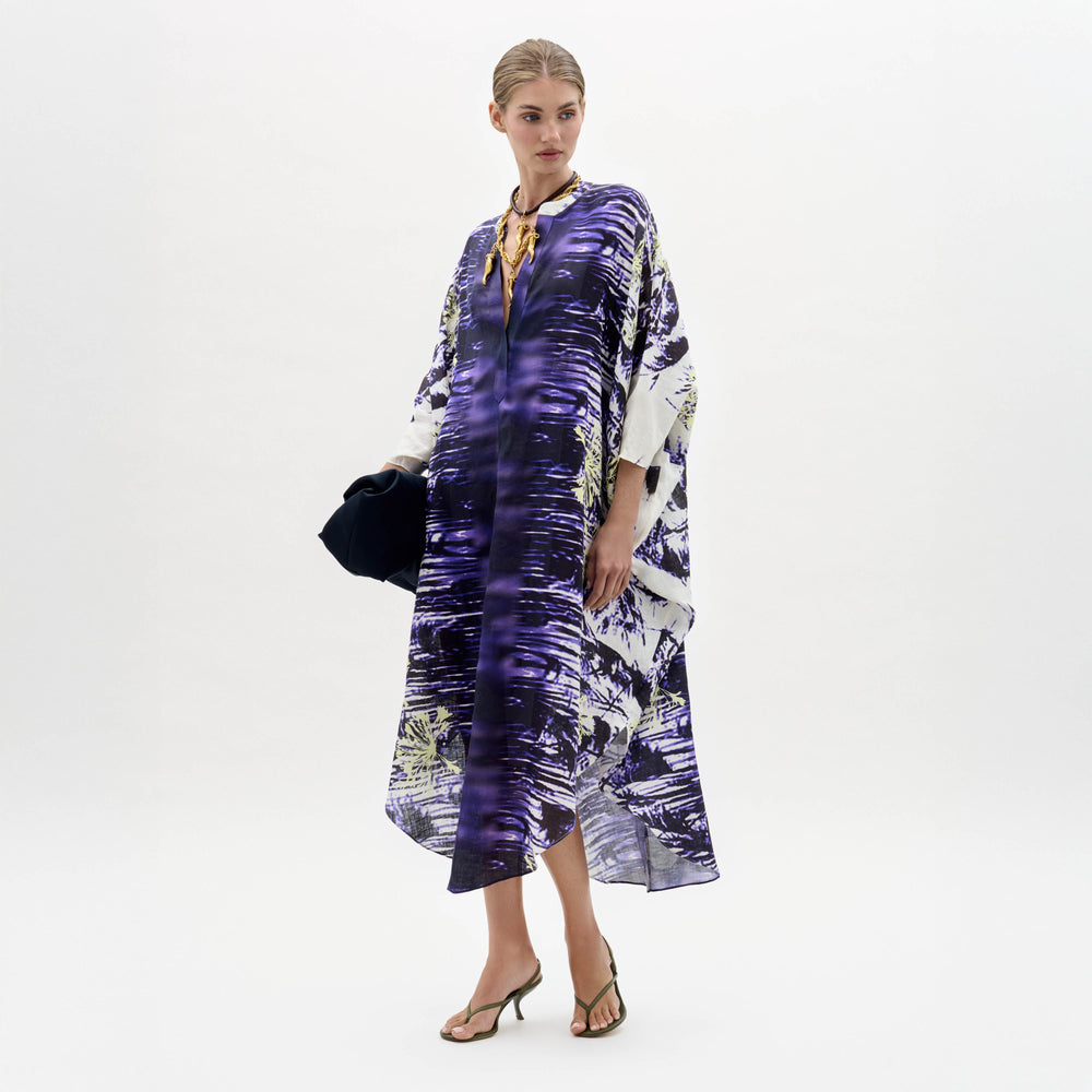 A woman stands against a plain white background wearing the Ilse Dress, a loose-fitting purple and white patterned piece, paired with a gold necklace, dark clutch, and heeled sandals.