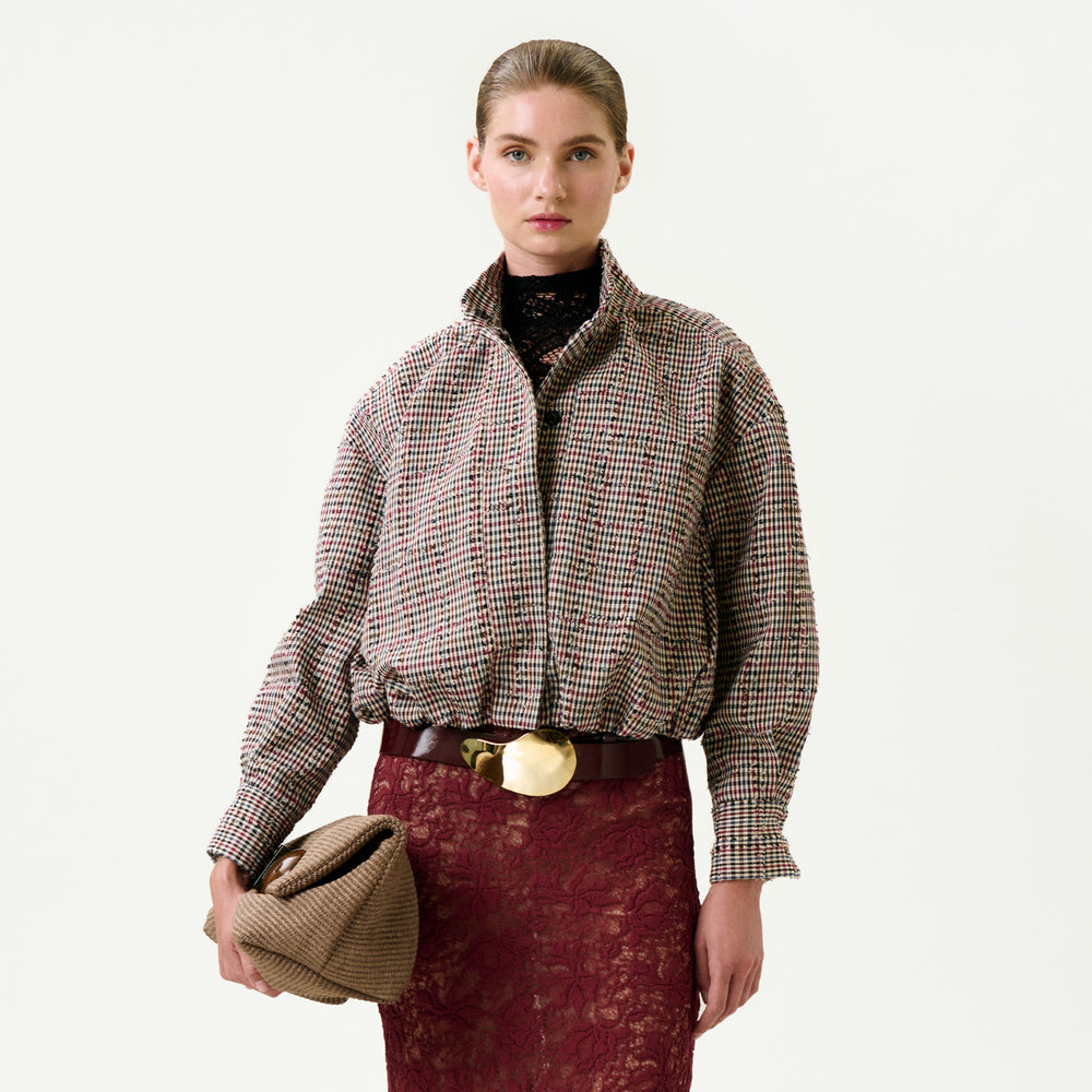 A woman stands against a white background, wearing the Julinka Jacket from Colombia in an oversized fit, paired with a dark lace skirt, a statement belt with a gold buckle, and holding a textured clutch bag. Her hair is pulled back neatly.