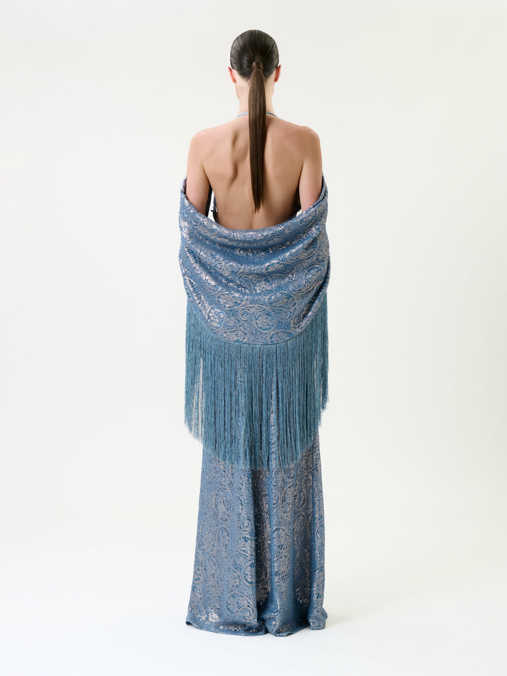 A person stands facing away, wearing the Habana Dress—a long, backless blue gown with an intricate pattern—and a matching fringed shawl draped over their shoulders. Their straight hair is tied back in a low ponytail.