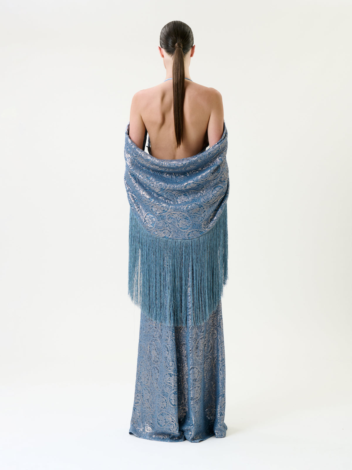 A person stands facing away, wearing the Habana Dress—a long, backless blue gown with an intricate pattern—and a matching fringed shawl draped over their shoulders. Their straight hair is tied back in a low ponytail.