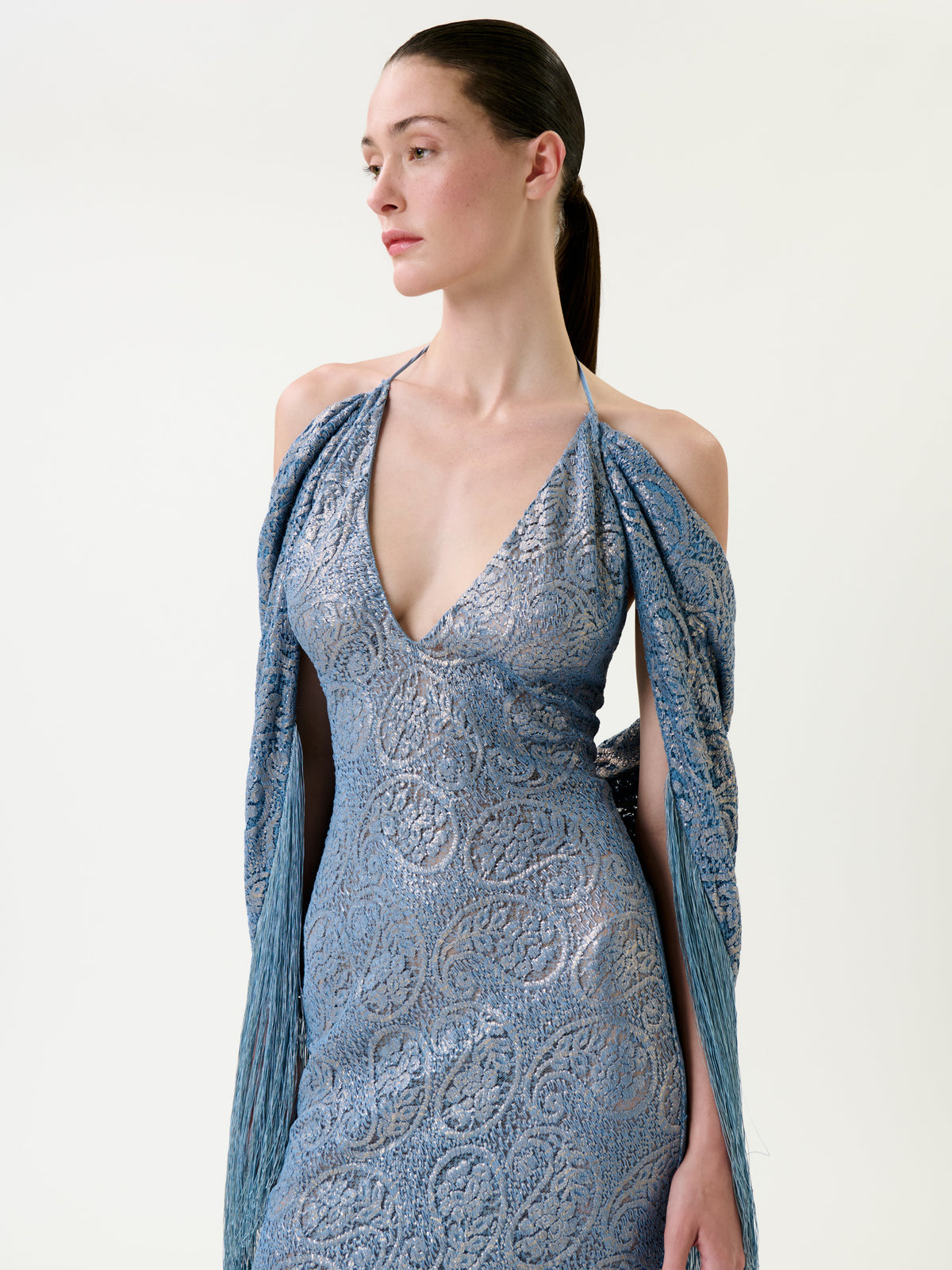 A woman with slicked-back hair wears the Habana Dress—a light blue, textured, sleeveless dress featuring a deep V-neck and draped shawl-like sleeves—as she stands against a plain white background, looking to the side.