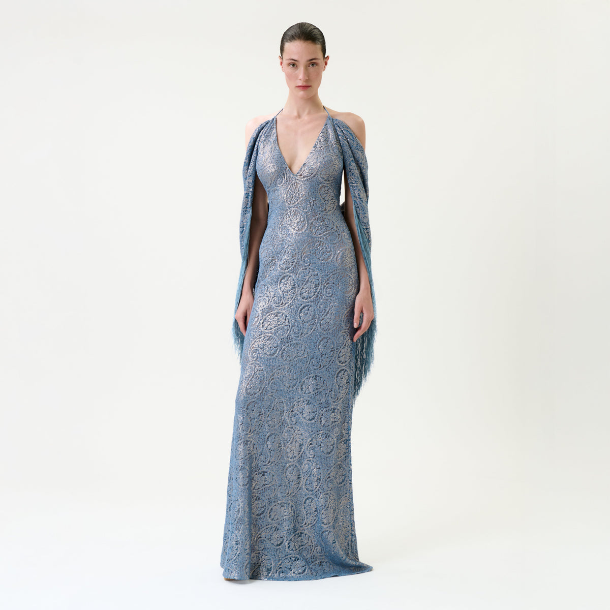 A woman poses against a simple backdrop wearing the Habana Dress, a floor-length, sleeveless blue lace gown featuring a halter neckline. She accessorizes with a matching shawl draped over her arms and has her hair slicked back.