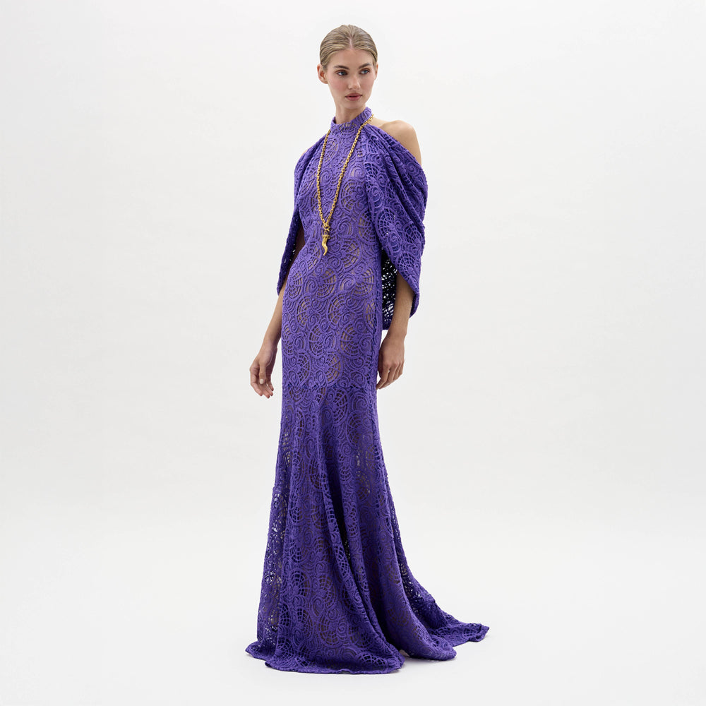 A woman models the Salvina Dress, a floor-length purple lace gown with cape detail, standing against a plain white background.
