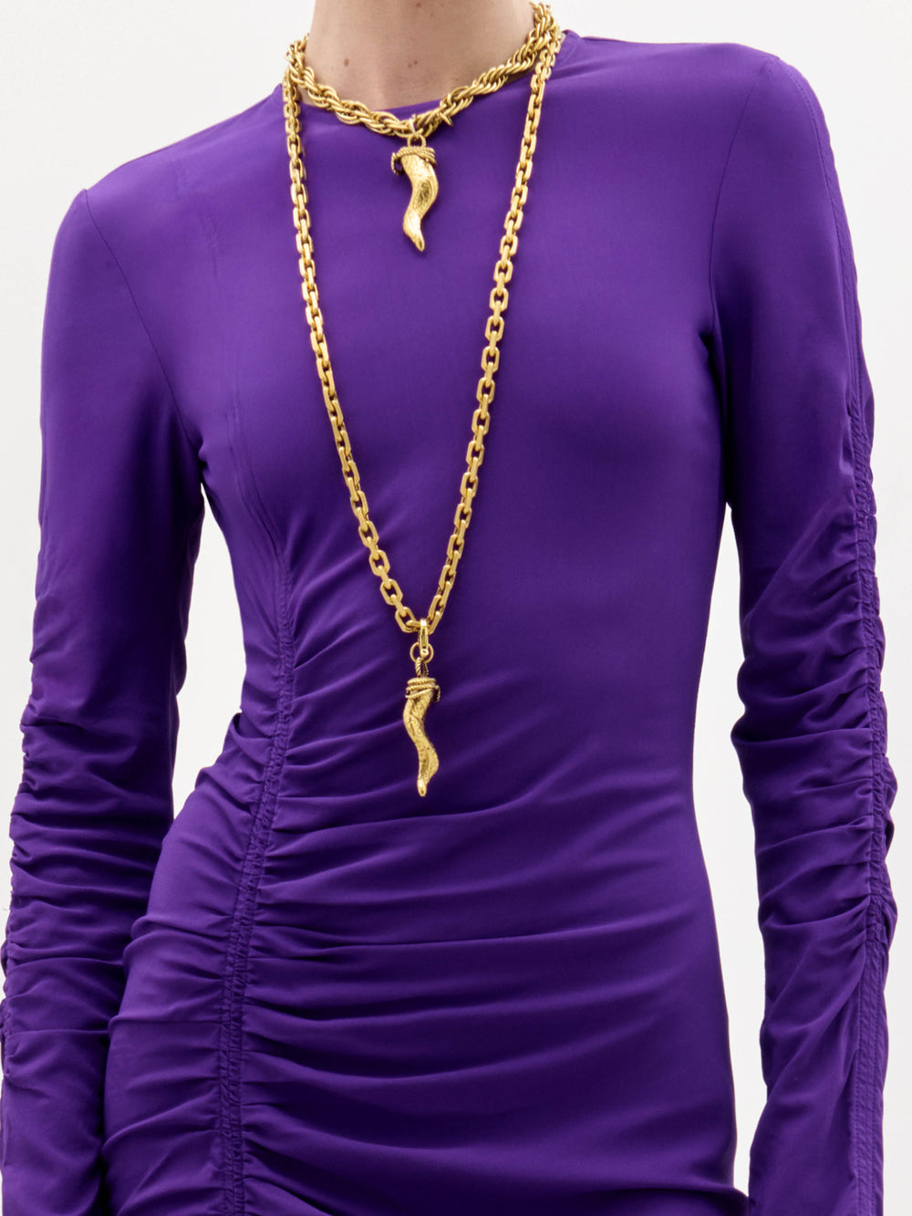 A woman stands against a plain white background, wearing the Mara Dress—a long-sleeved, asymmetrical purple dress with ruching—paired with black sandals and a long gold necklace.
