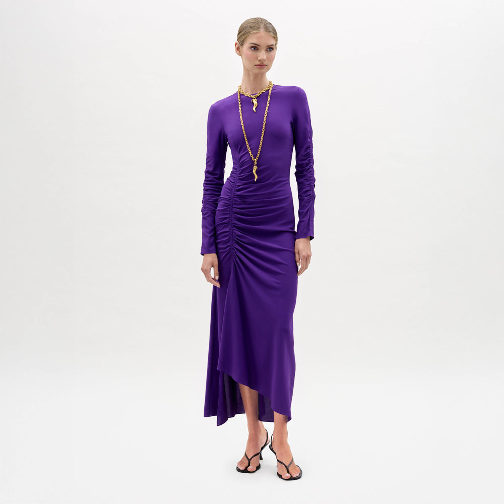 A woman stands against a plain white background, wearing the Mara Dress—a long-sleeved, asymmetrical purple dress with ruching—paired with black sandals and a long gold necklace.