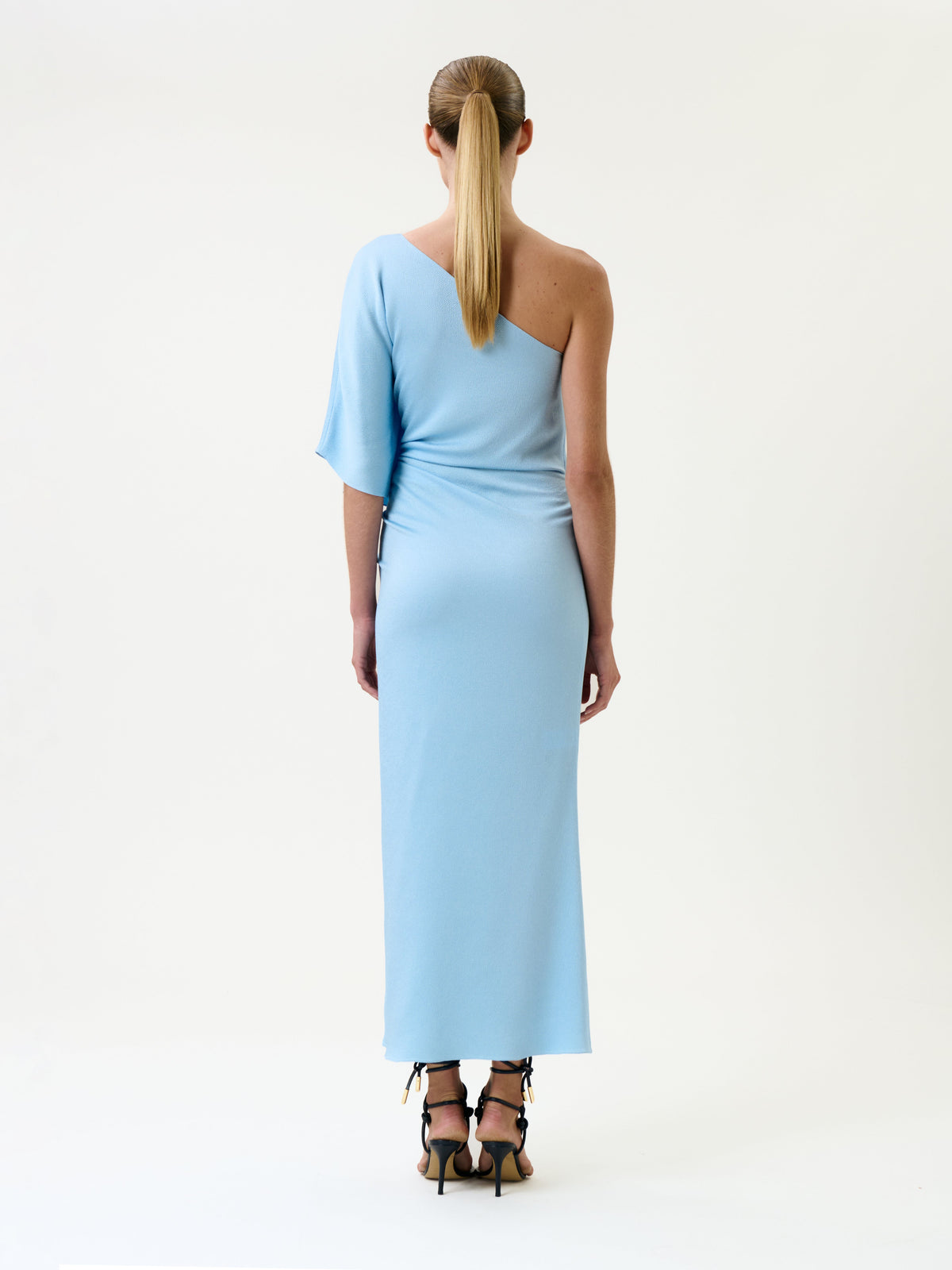 A person with long, light brown hair in a ponytail wears the Oby Dress—a light blue, one-shoulder, ankle-length dress—paired with black heels, standing against a plain white background and facing away from the camera.