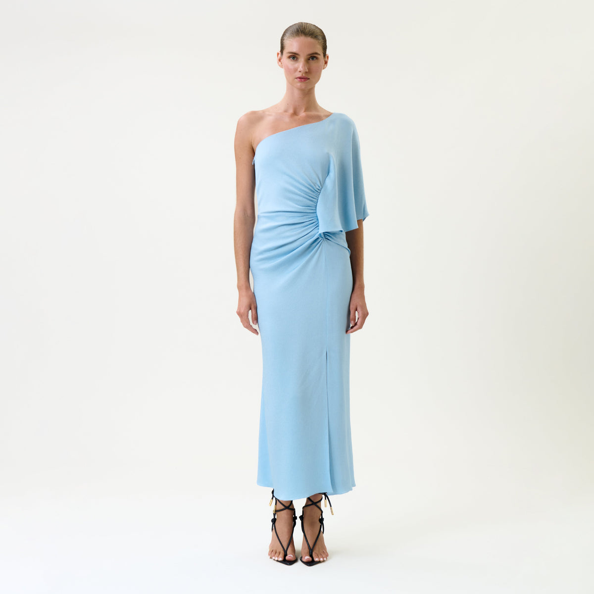 A woman stands against a plain white background wearing the Oby Dress, a light blue, one-shoulder, ruched midi dress, paired with black strappy heels.