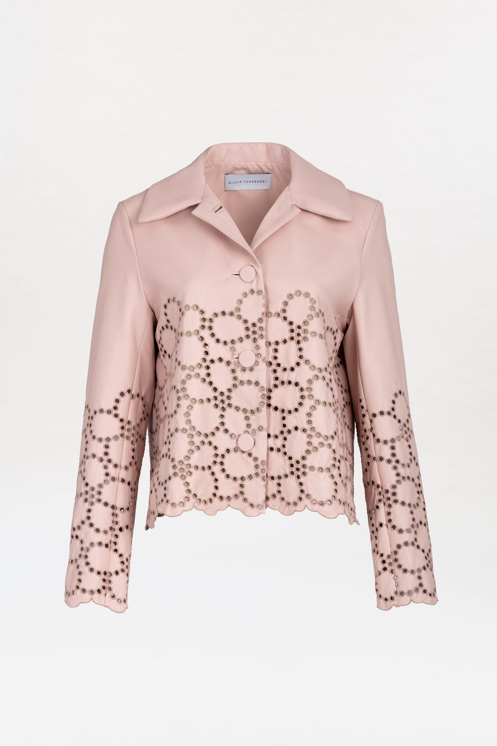 The Belen Jacket is a light pink, collared jacket featuring a scalloped hem and circular cut-out patterns on the lower half and sleeves. It has a button closure and boasts a smooth, tailored design.