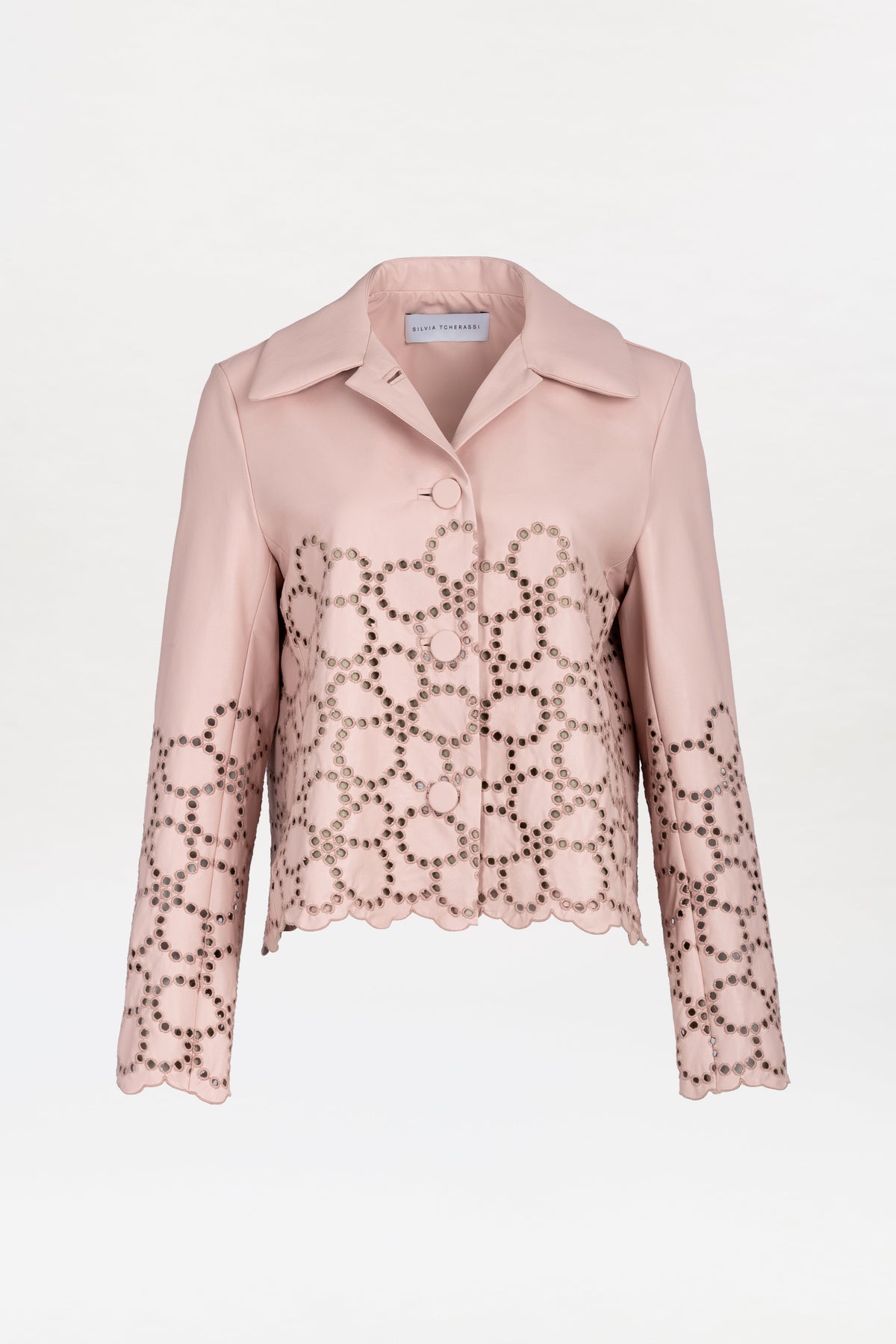 The Belen Jacket is a light pink, collared jacket featuring a scalloped hem and circular cut-out patterns on the lower half and sleeves. It has a button closure and boasts a smooth, tailored design.