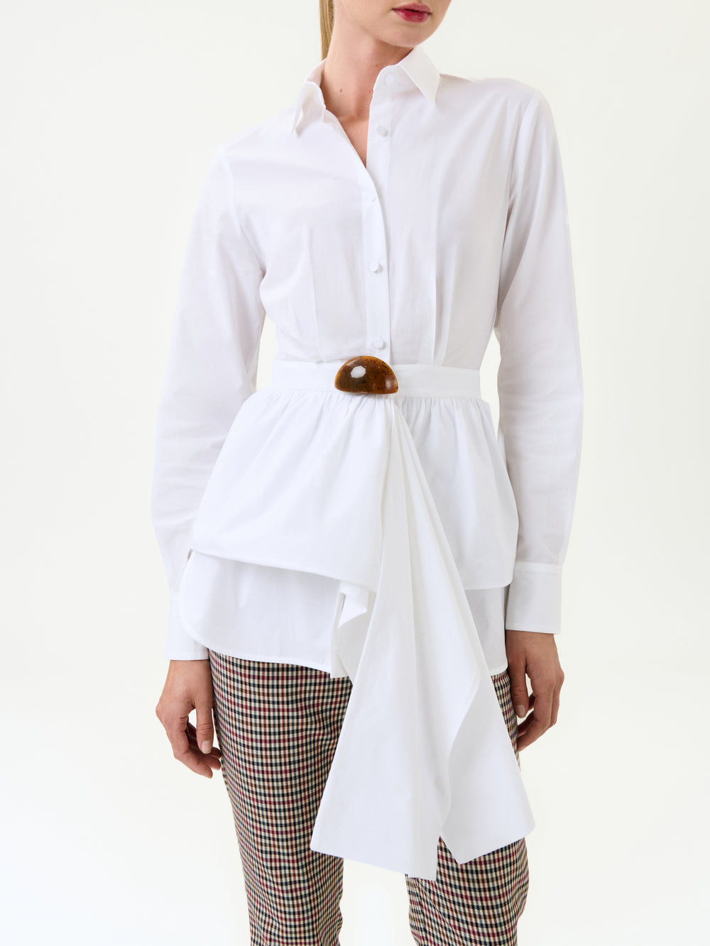A woman wears the Malina Shirt, a white long-sleeve button-up with an asymmetric layered design, made from Portuguese fabric and finished with a wide belt featuring a brown decorative buckle, paired with plaid pants.