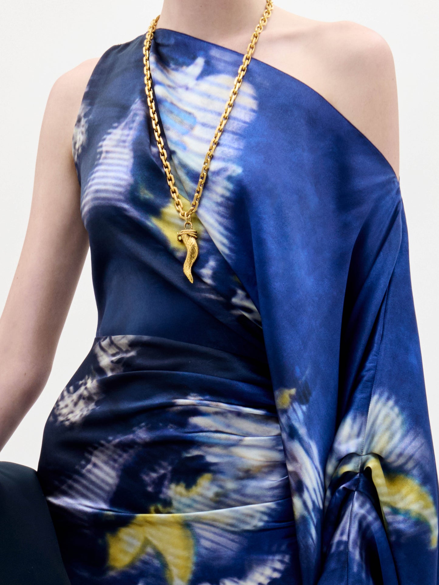 A woman stands against a plain background wearing the Dotty Dress, a one-shoulder blue dress with a yellow and white floral pattern, and holds a black clutch with a fringe strap.