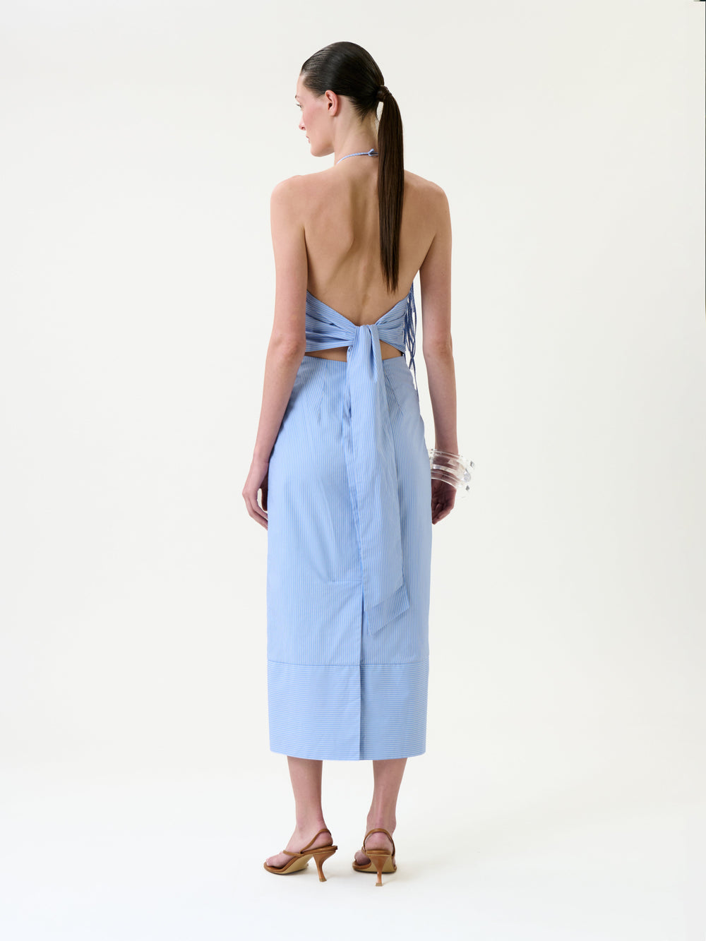 A woman stands with her back to the camera, wearing the Otilia Dress—a light blue, backless halter style with a bow detail and a mid-length skirt—paired with brown heeled sandals and clear bracelets. Her straight hair is tied in a low ponytail, set against a plain white background.
