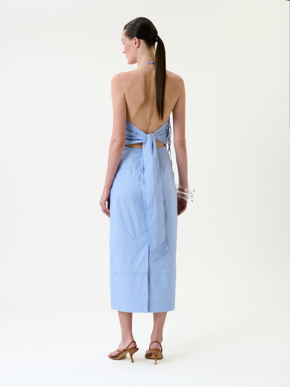 A woman stands with her back to the camera, wearing the Otilia Dress—a light blue, backless halter style with a bow detail and a mid-length skirt—paired with brown heeled sandals and clear bracelets. Her straight hair is tied in a low ponytail, set against a plain white background.