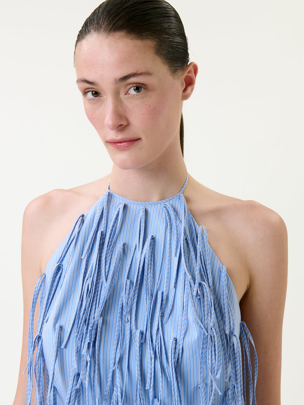 A woman with straight brown hair pulled back wears the Otilia Dress, a sleeveless halter-neck style featuring blue vertical stripes and long, fringed fabric details on the front. She is looking at the camera against a plain light background.