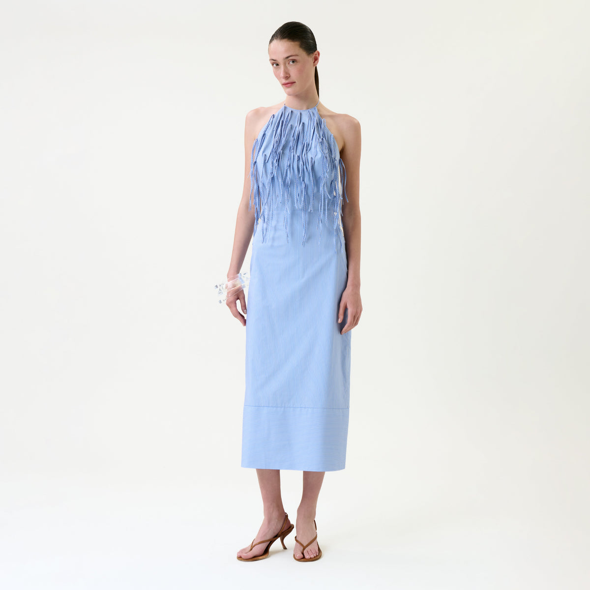 A woman stands against a plain background wearing the Otilia Dress, a light blue sleeveless midi dress featuring fringe detailing on the front, paired with brown heeled sandals. Her hair is tied back and she accessorizes with a white bracelet.