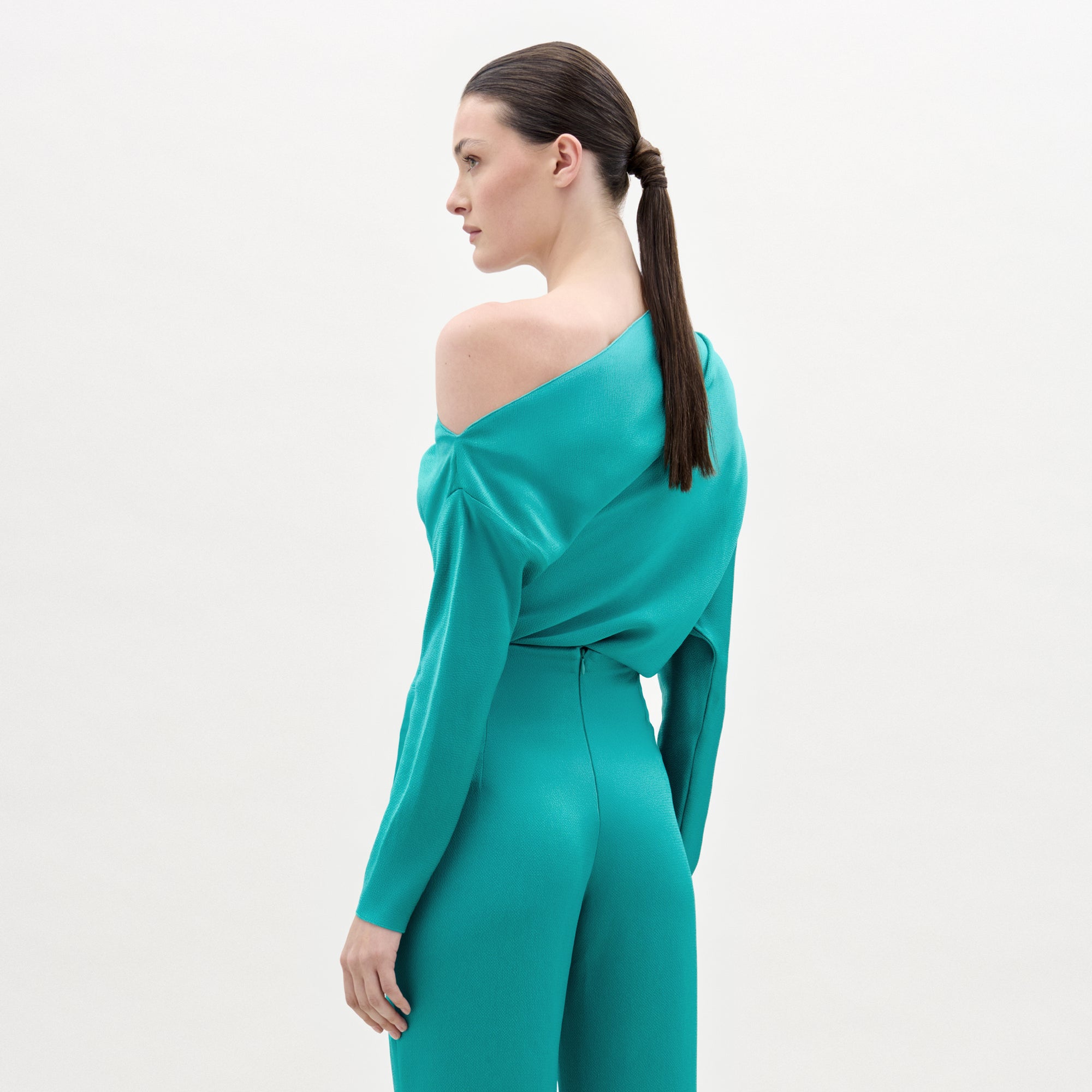 A woman with straight brown hair in a low ponytail, wearing the turquoise Viola Blouse made from Italian fabric, stands sideways against a plain light background.