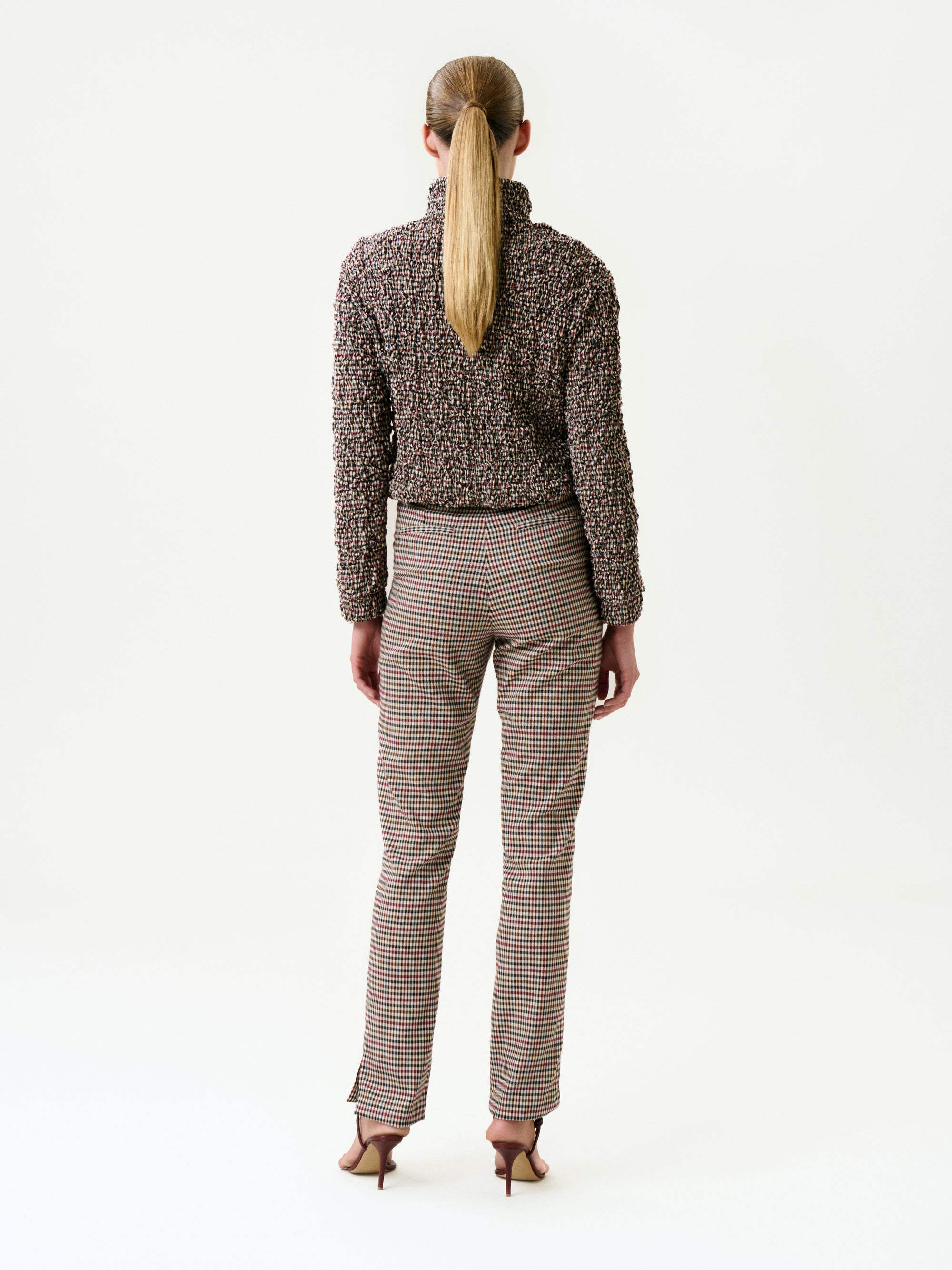 A person with long straight hair in a low ponytail stands facing away, wearing the Luca Pant, a textured long-sleeve Italian-fabric top, and heeled sandals against a plain white background.