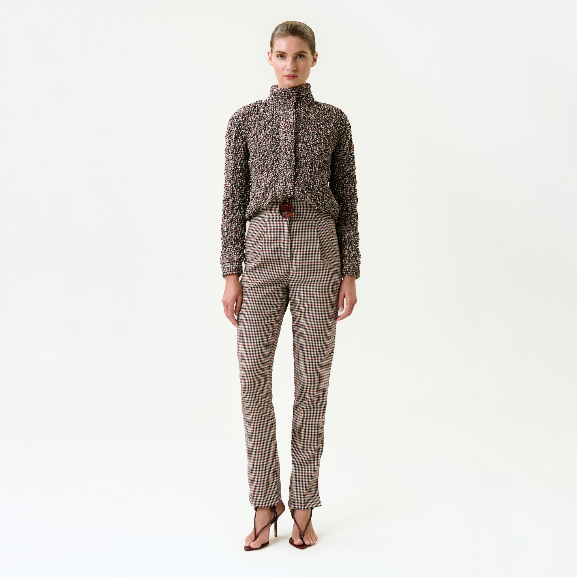 A woman stands against a white background wearing the Luca Pant—high-waisted, belted plaid pants made from Italian fabric for a true-to-size fit—with a textured long-sleeve top and open-toed heels; her hair is pulled back.