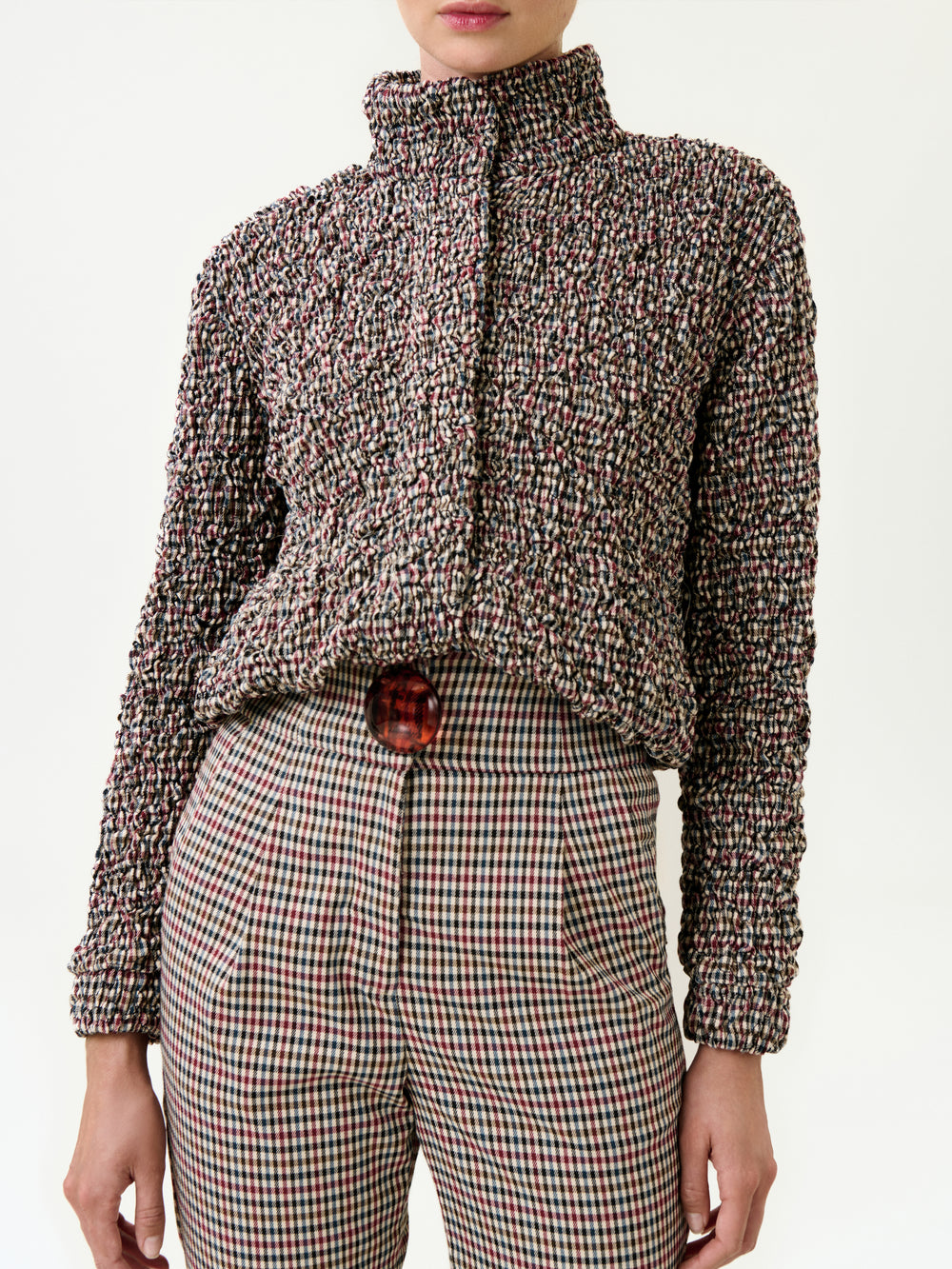 A person wearing the Zalta Jacket, a relaxed fit piece made in Colombia, over a textured multicolored knit sweater and high-waisted plaid pants with a large red button, stands against a plain light background.