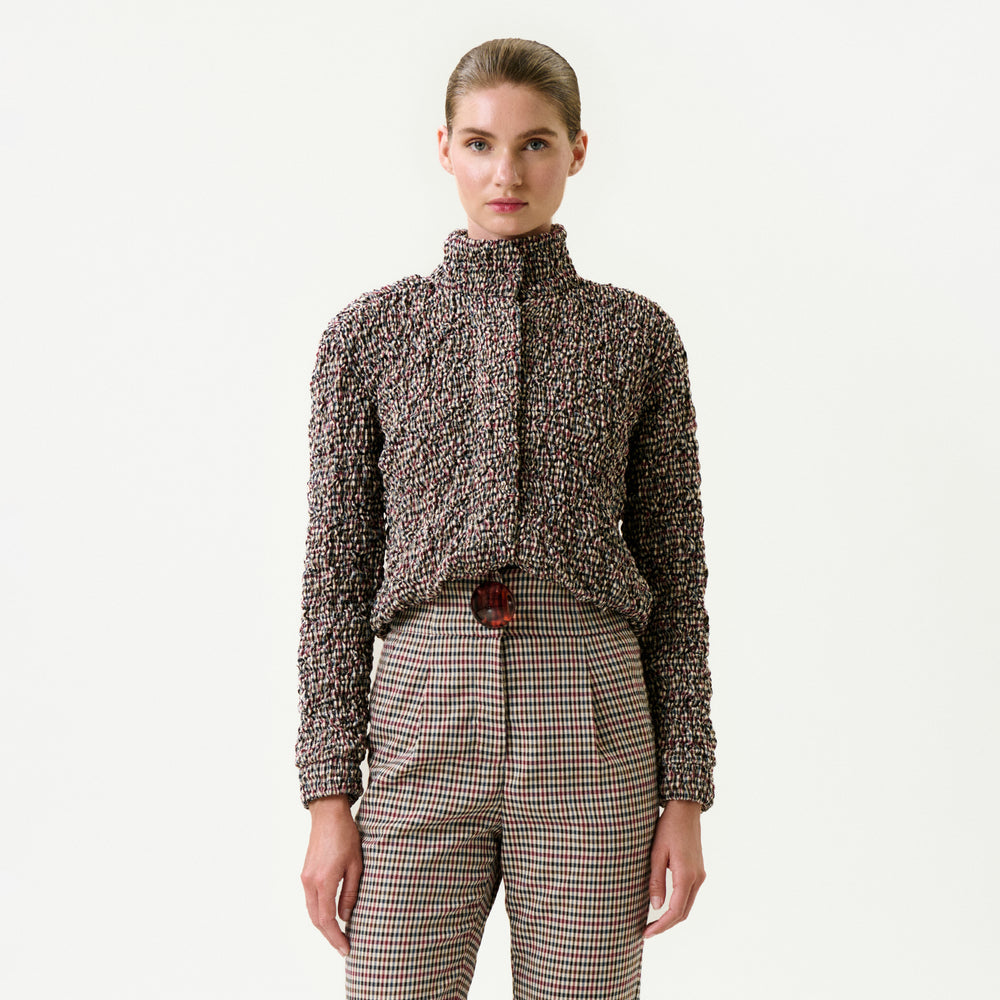 A woman stands against a plain white background, wearing the Zalta Jacket in a speckled pattern and high-waisted checked trousers with a red belt, creating a modern and effortless look.