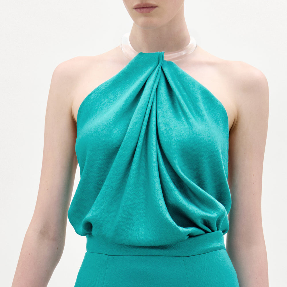 A person wears the Dianela Blouse, a sleeveless teal halter-top with a draped front in luxurious Italian fabric, paired with matching high-waisted pants and a translucent choker necklace, standing against a plain white background.