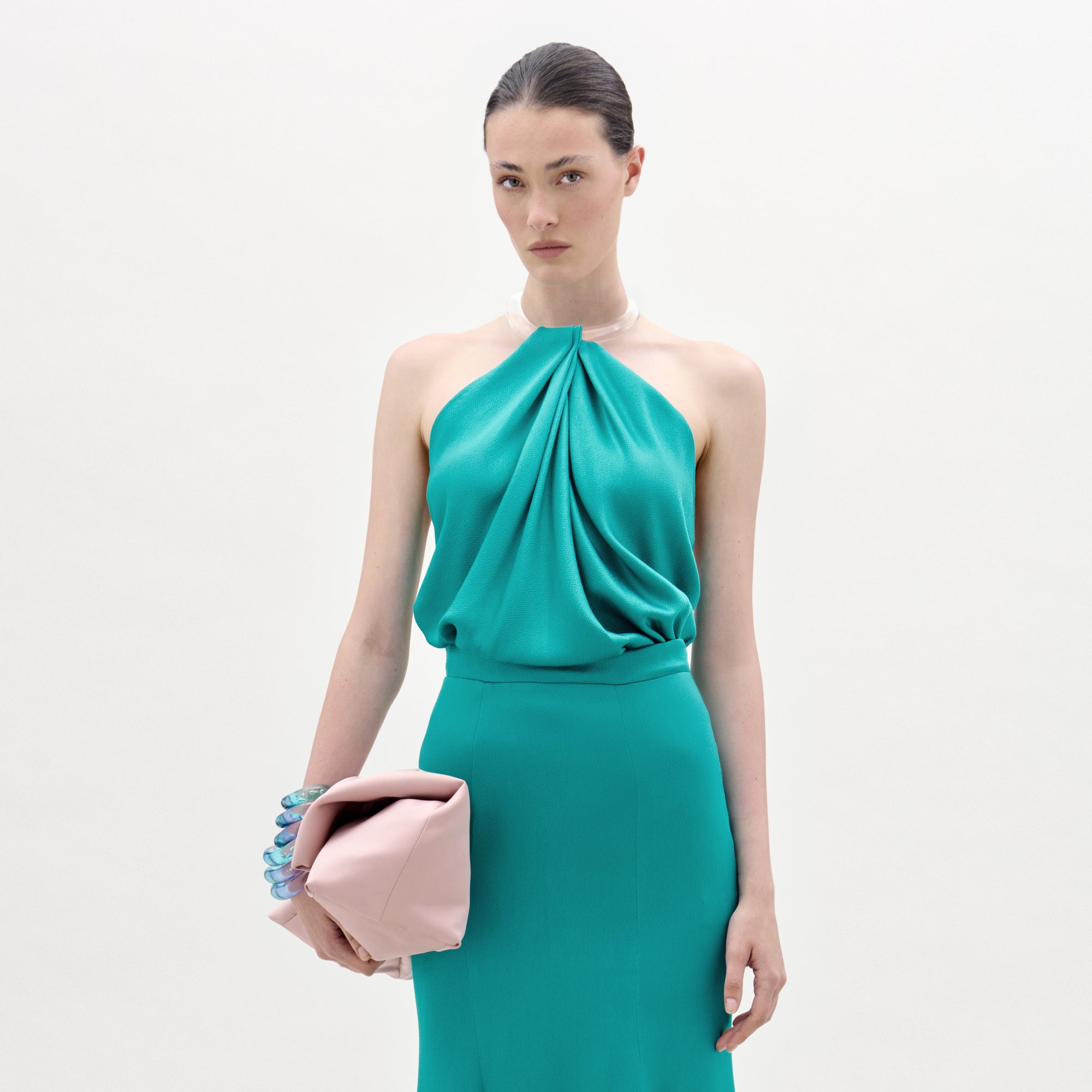 A woman in a Dianela Blouse poses against a plain white background, holding a large pale pink clutch and wearing several chunky rings, looking directly at the camera with a neutral expression.