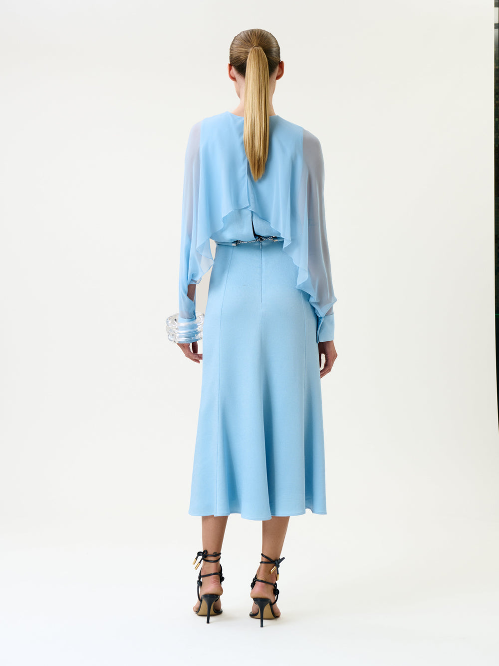 A woman stands facing away, wearing the Rubina Dress—a light blue design featuring sheer long sleeves and ruffled back detail. She has a ponytail and wears black strappy high-heeled sandals. The background is plain white.