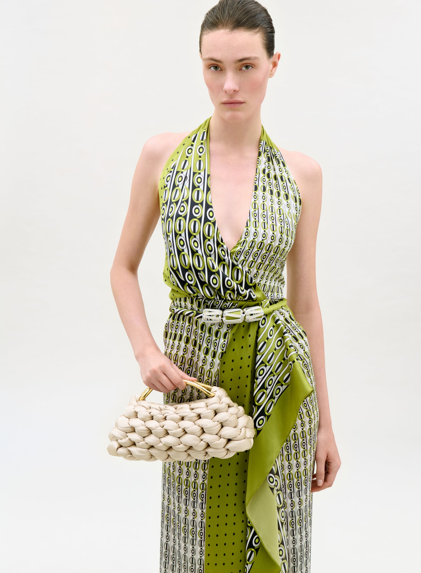 Woman wearing a green and white patterned dress holding a woven handbag on a white background