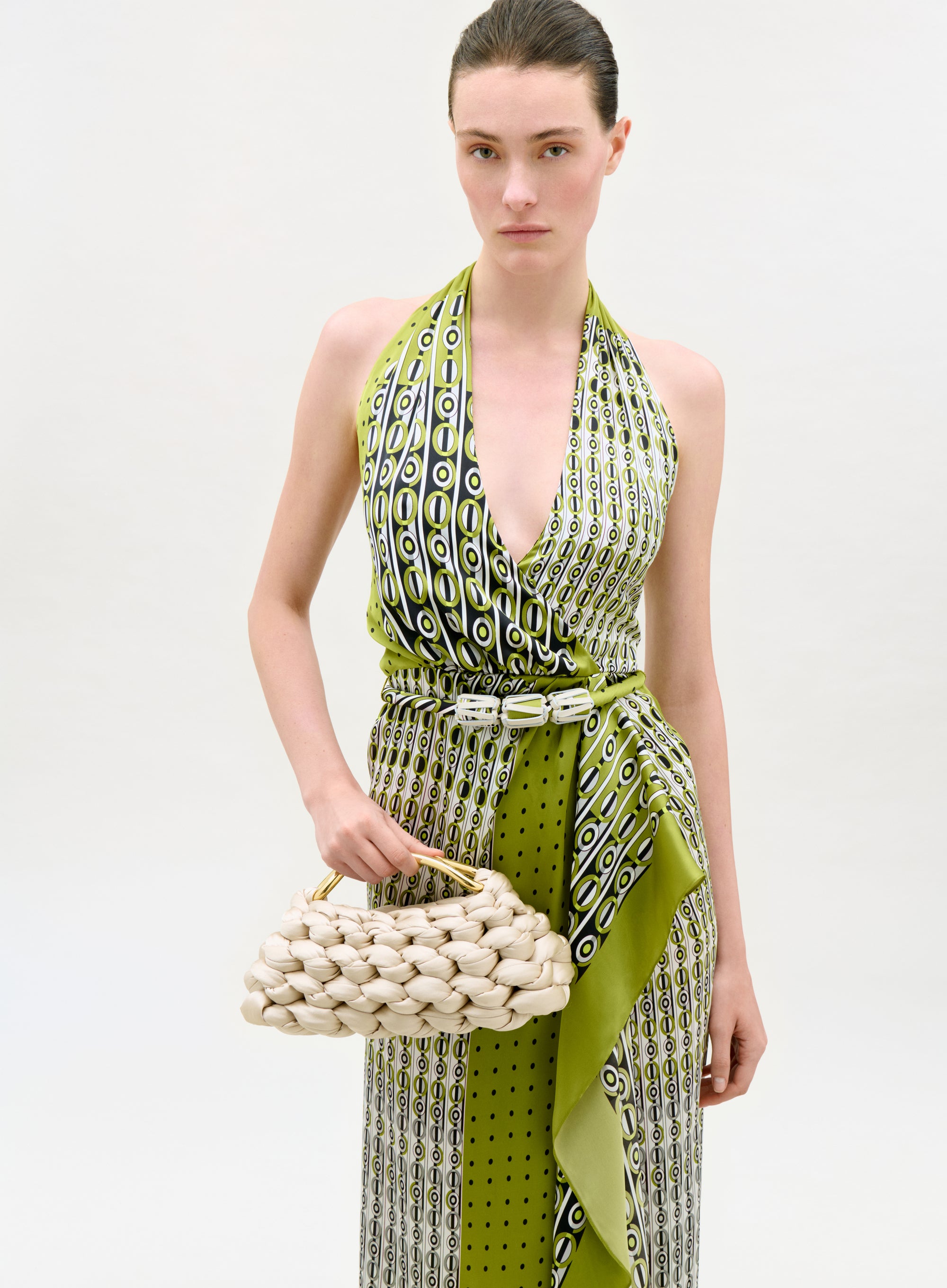 Woman wearing a green and white patterned dress holding a woven handbag on a white background