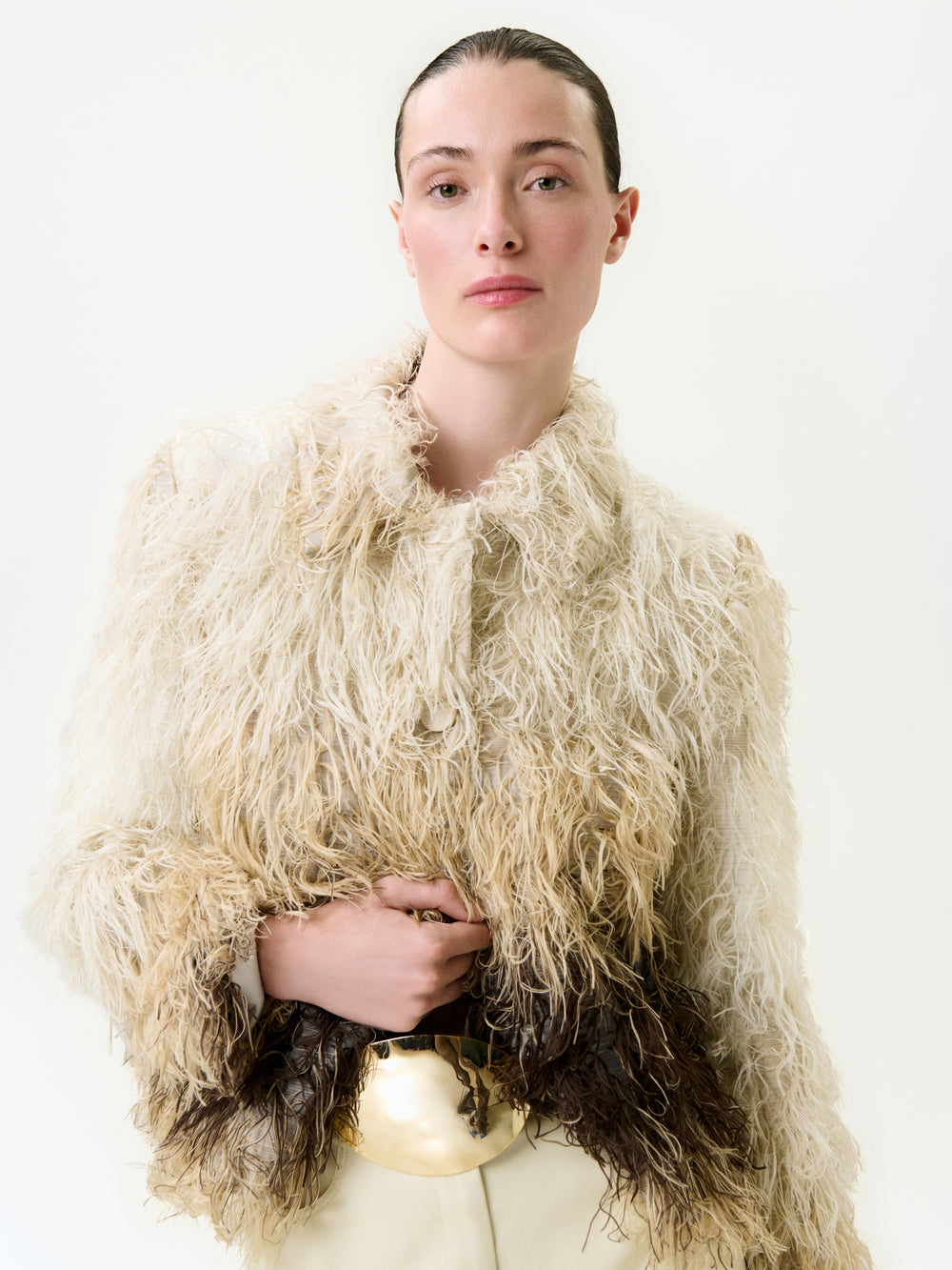 A person with slicked-back hair wears the Columba Jacket, a textured, shaggy off-white and brown Italian fabric piece with a true-to-size fit, paired with cream trousers and a large gold belt buckle against a plain light background.