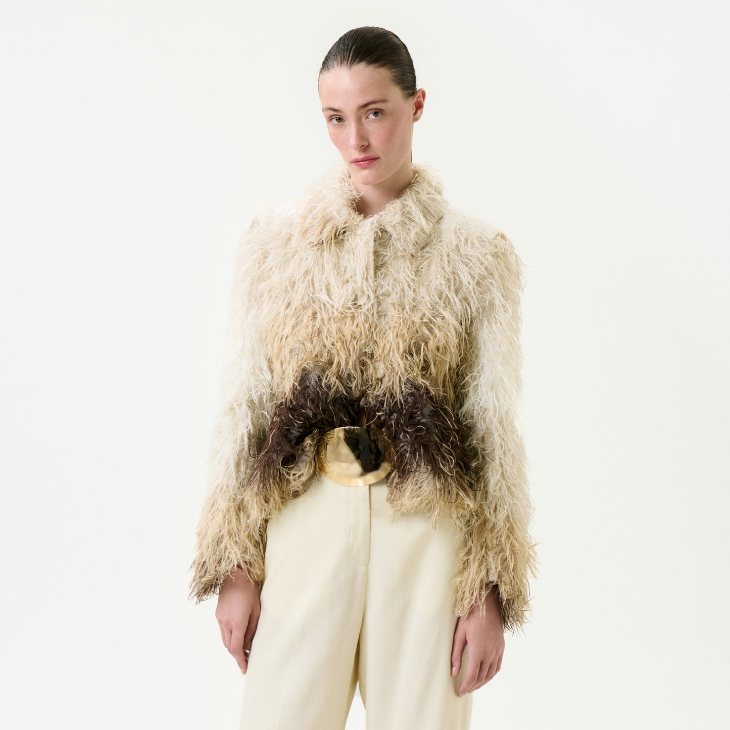 A person with slicked-back hair wears the Columba Jacket, a textured, shaggy cream and brown fur Italian fabric piece, paired with high-waisted cream pants, standing against a plain white background.