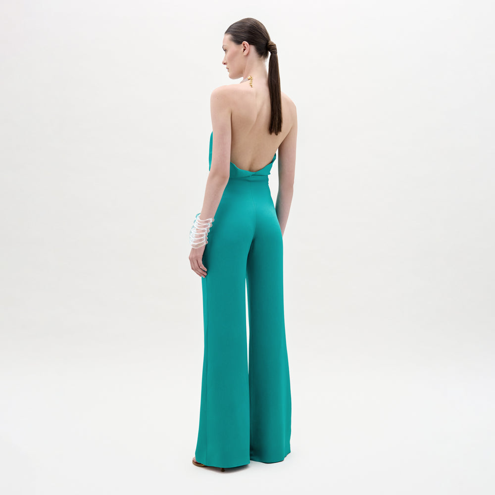 A woman faces away, wearing the teal Palermo Pant, a backless halter-neck jumpsuit with wide-leg pants made from Italian fabric. Her hair is tied back and she accessorizes with white bangles against a pale background.