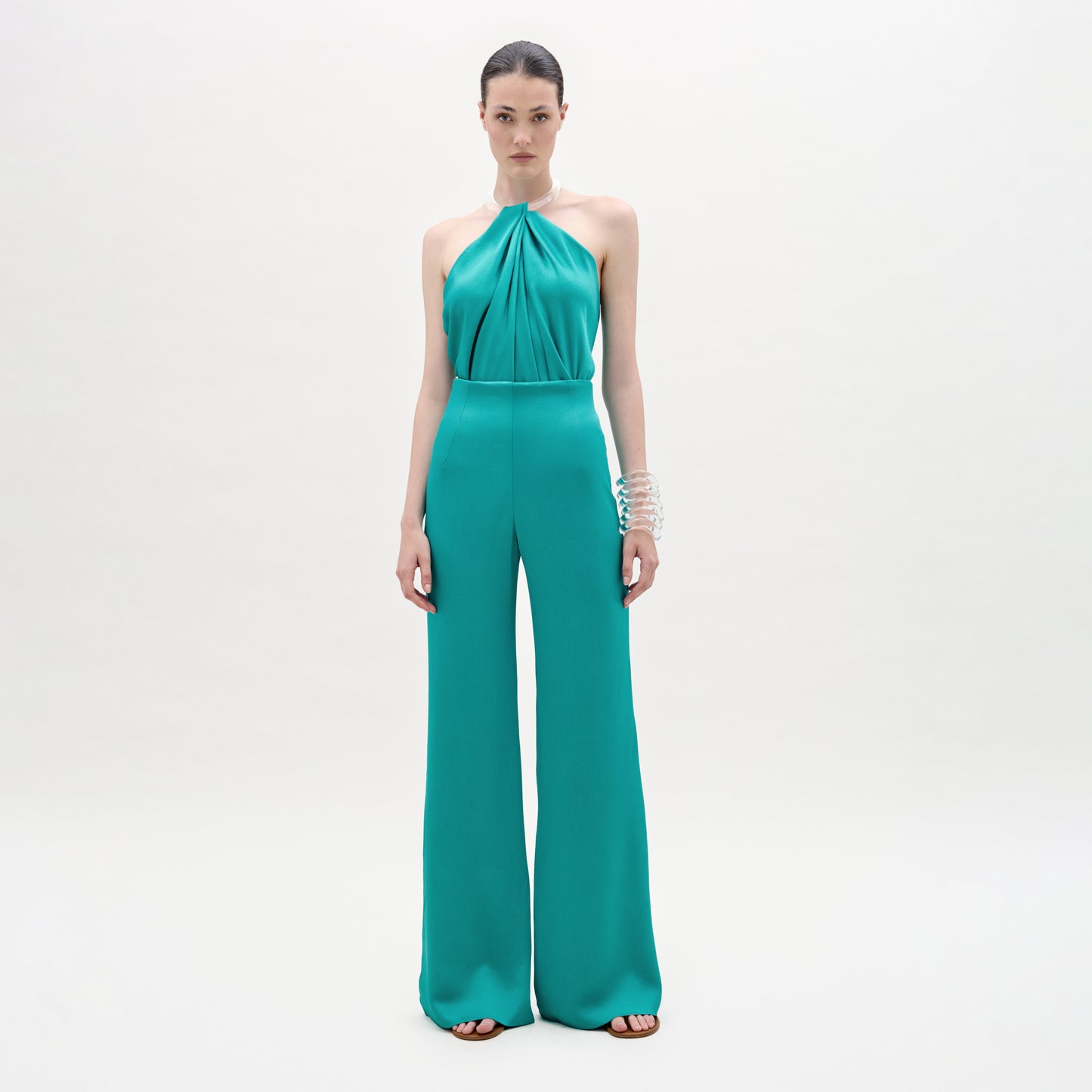 A woman stands against a light background wearing the Palermo Pant—a teal, wide-leg, Italian-fabric jumpsuit with a halter neck. Her hair is slicked back, and she accessorizes with stacked silver bracelets on her right arm.