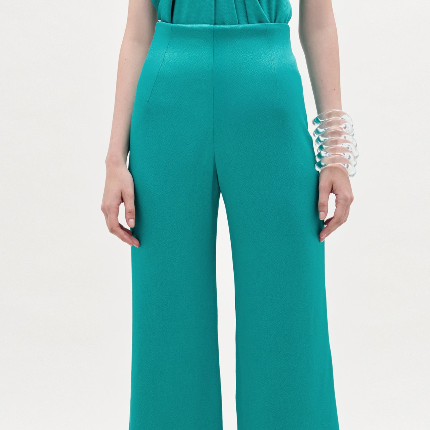 A person wearing the Palermo Pant, a high-waisted hammered satin wide-leg style in teal, with a matching top and a large clear chunky bracelet, stands against a plain light background.