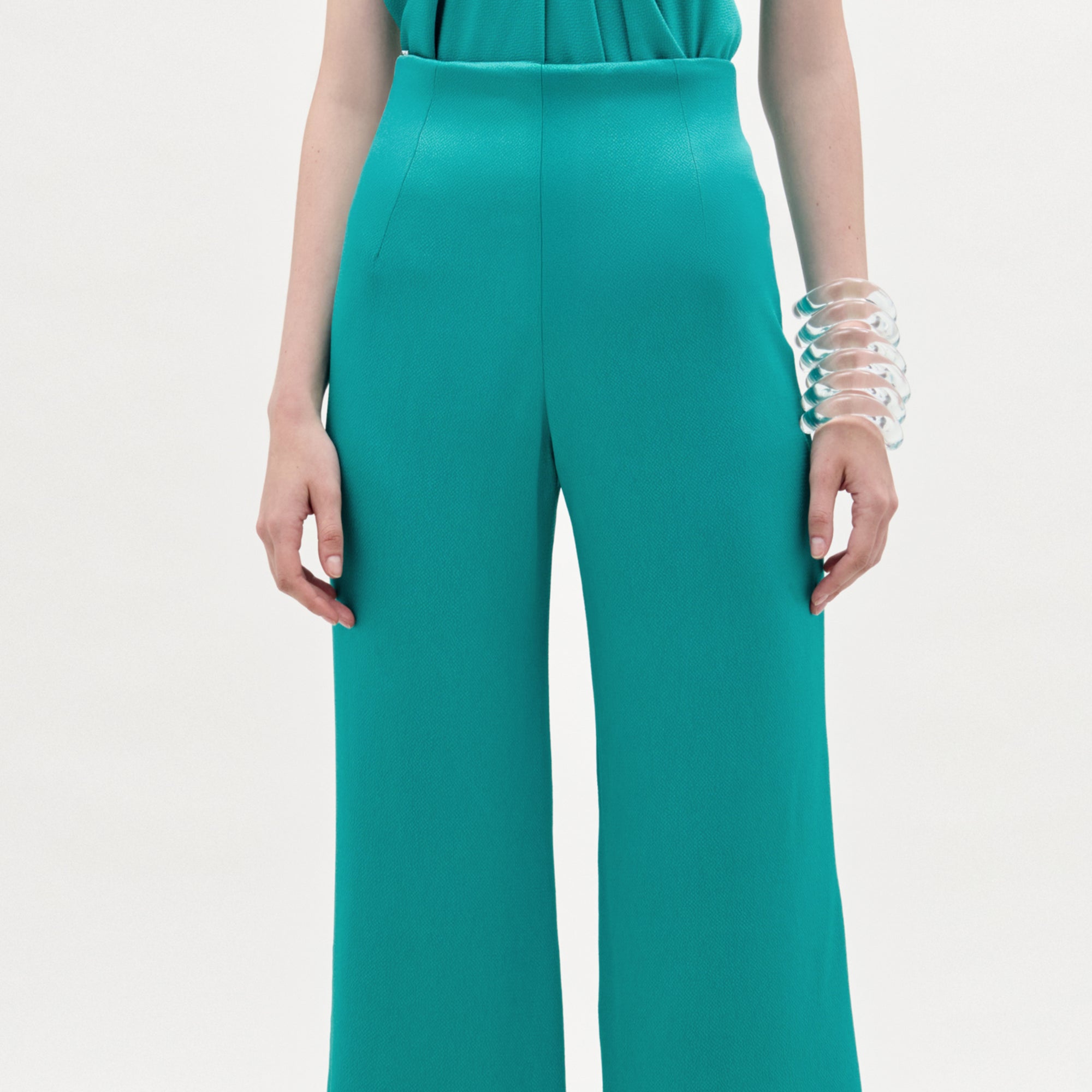 A person wearing the Palermo Pant, a high-waisted hammered satin wide-leg style in teal, with a matching top and a large clear chunky bracelet, stands against a plain light background.