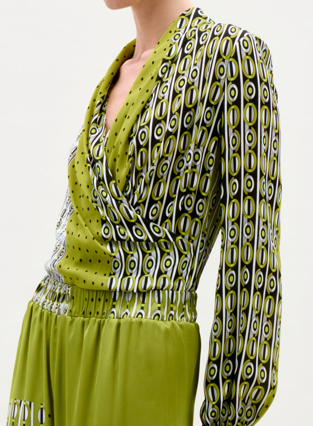 A person wearing the Tea Blouse—a green, long-sleeve top with a draped neckline, bold black, white, and yellow geometric oval patterns with small black dots. The solid green skirt complements the blouse.