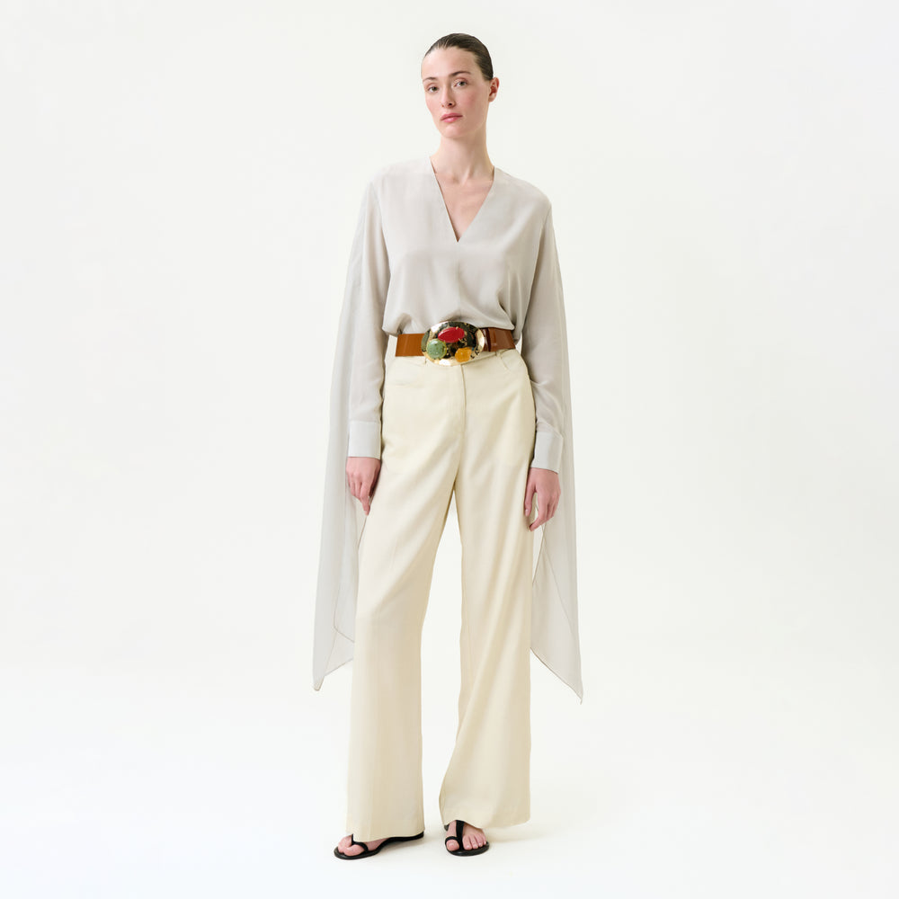 A woman stands against a white background wearing a light beige Italian-fabric blouse with draped sides, the Emine Pant—wide-leg cream trousers—a brown belt featuring a large floral buckle, and black sandals.