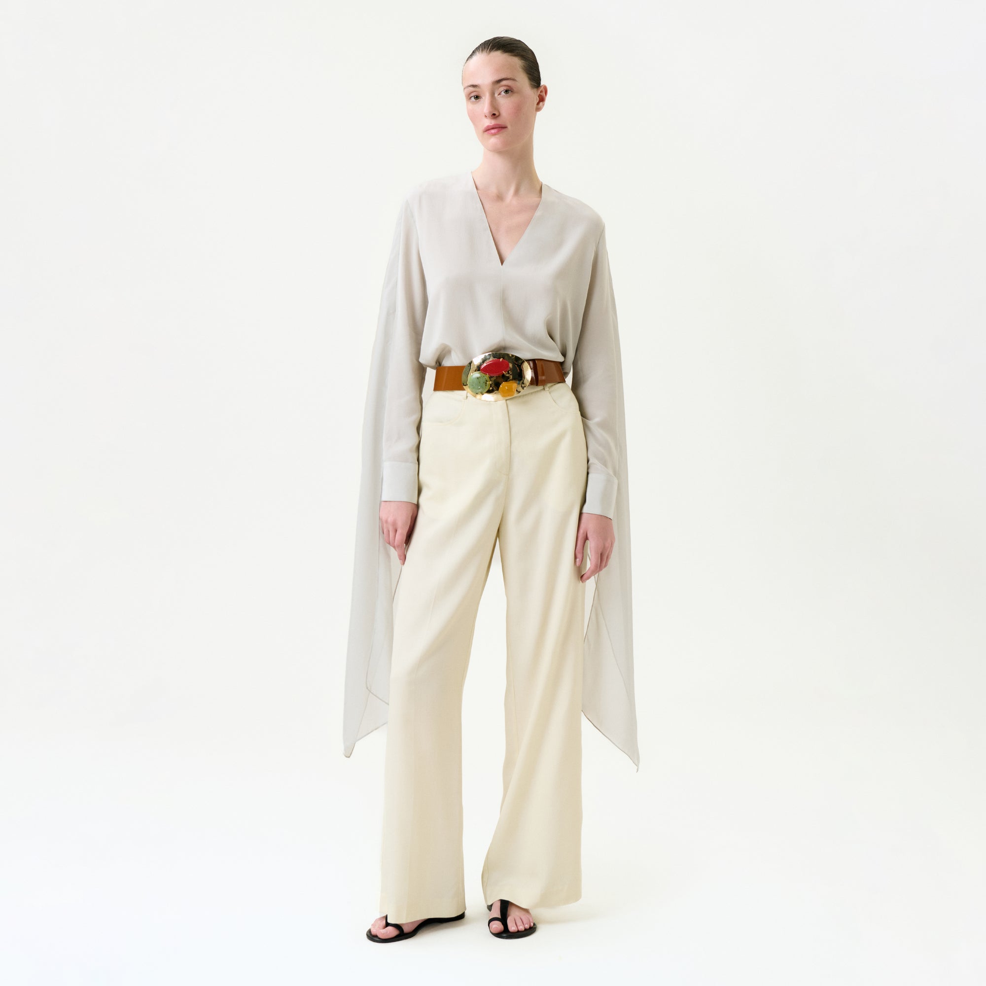 A woman stands against a white background wearing a light beige Italian-fabric blouse with draped sides, the Emine Pant—wide-leg cream trousers—a brown belt featuring a large floral buckle, and black sandals.
