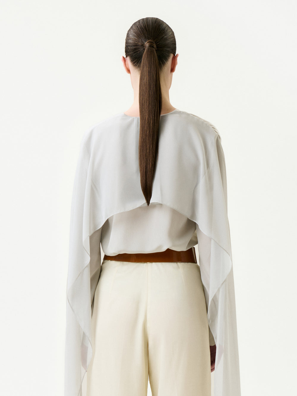 A person with long brown hair in a low ponytail is seen from the back wearing the Vanna Blouse, a light, flowy, sheer top crafted from 100% silk Italian fabric with wide sleeves, paired with cream pants against a plain white background.
