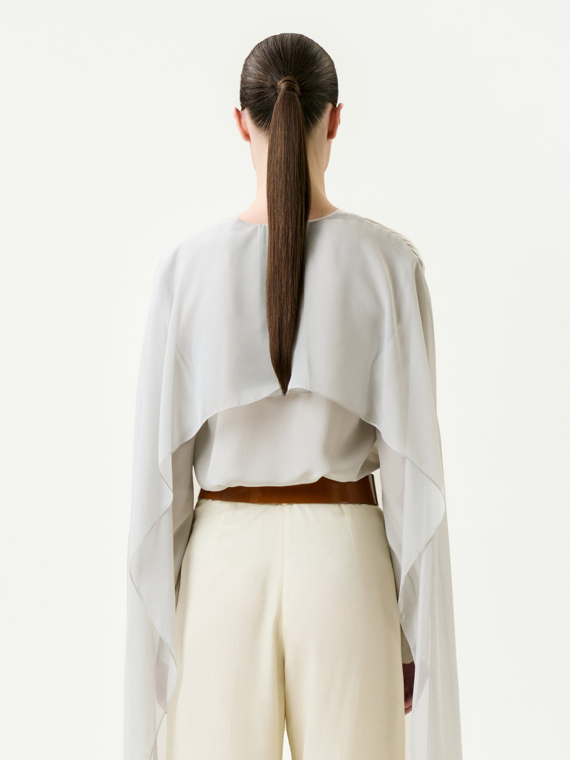 A person with long brown hair in a low ponytail is seen from the back wearing the Vanna Blouse, a light, flowy, sheer top crafted from 100% silk Italian fabric with wide sleeves, paired with cream pants against a plain white background.