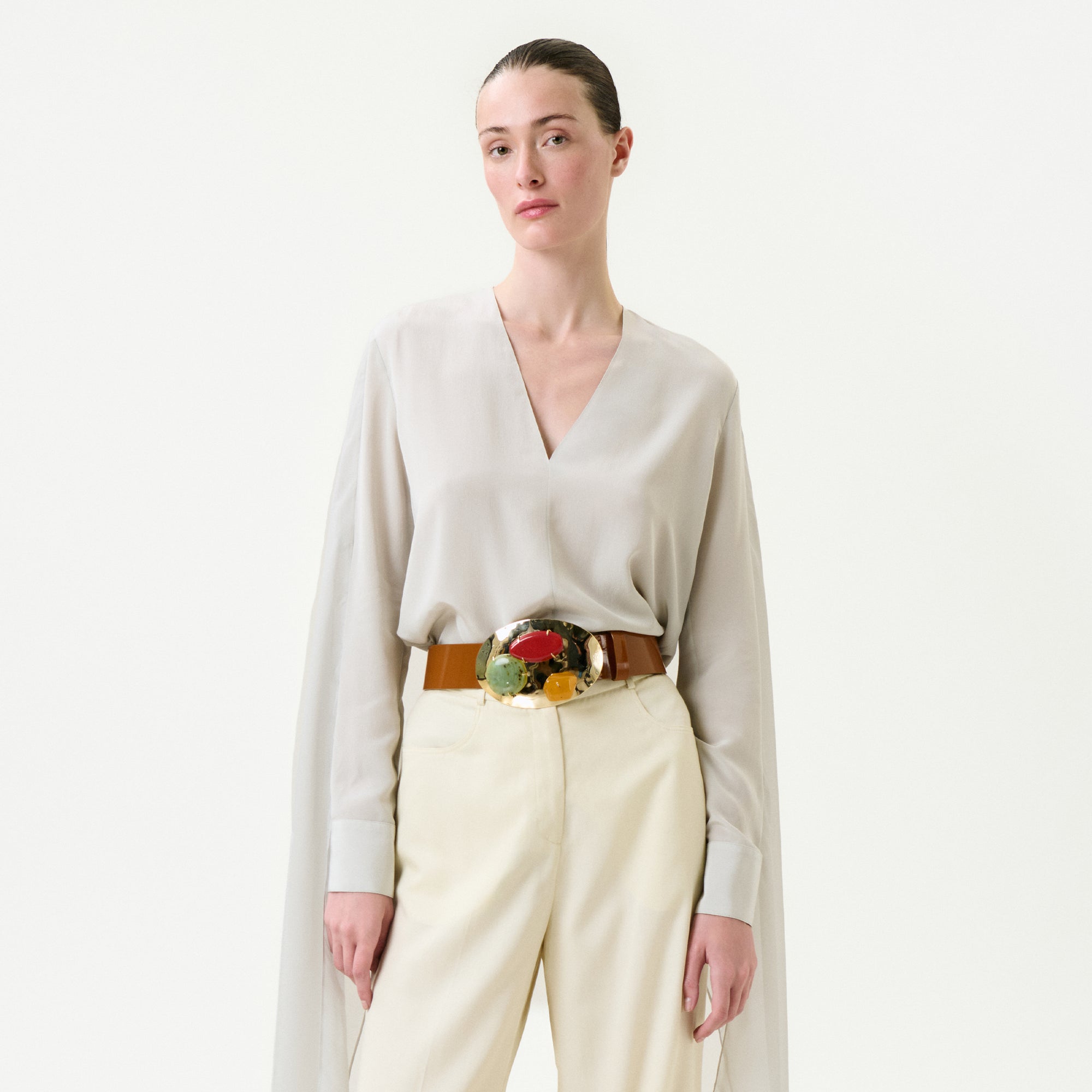 A woman stands against a white background wearing the Vanna Blouse in light beige Italian fabric with wide sleeves, paired with cream high-waisted pants and a brown belt featuring a large, colorful decorative buckle.
