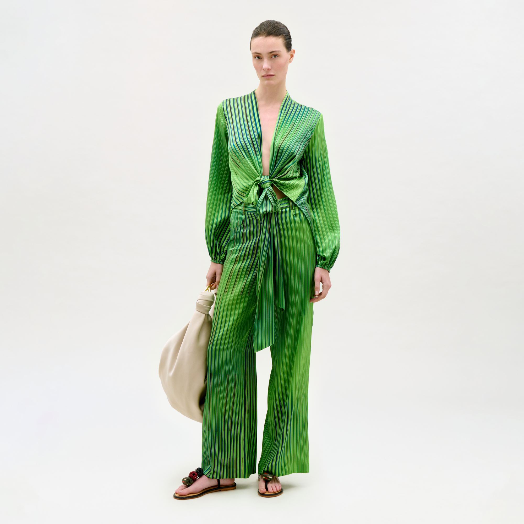 Woman wearing a green pleated outfit holding a beige handbag on a white background