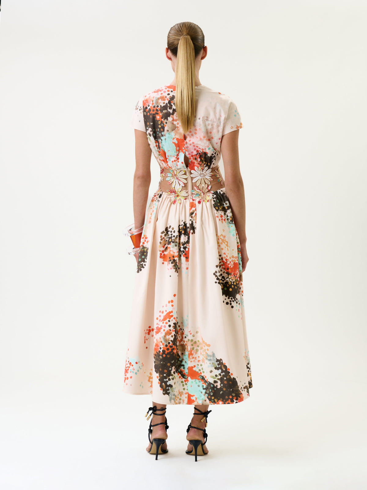 A woman with a ponytail wears the Macaria Dress, a light-colored, short-sleeve, ankle-length piece featuring a colorful splatter pattern. She stands facing away to reveal the dress’s gathered waist and pairs it with strappy high heels.