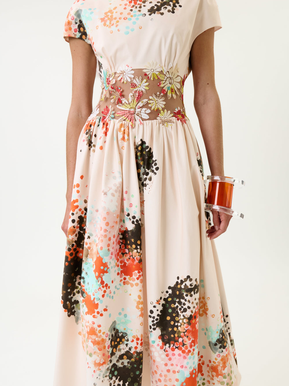 A woman wearing the Macaria Dress—a cream dress featuring a colorful paint splatter pattern and a sheer floral waistband—holds a transparent orange clutch. The image is cropped at the shoulders and mid-calf.