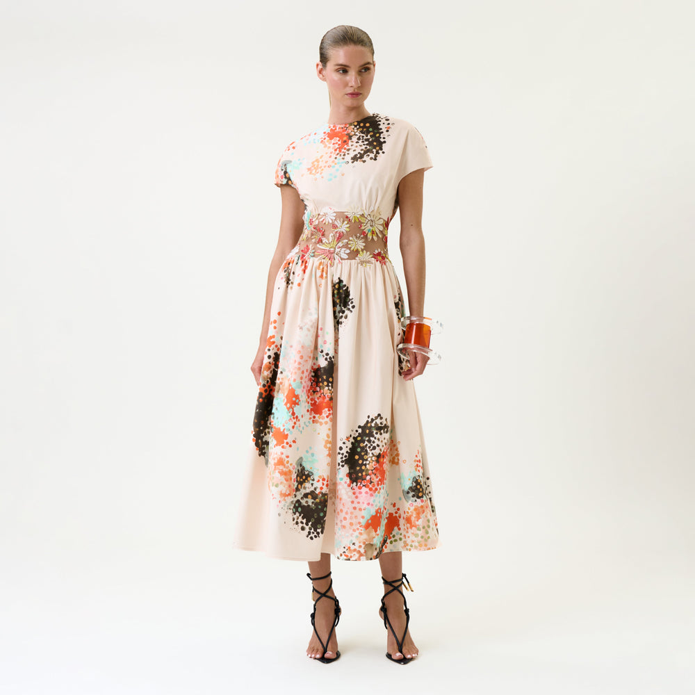A woman stands against a plain white background wearing the Macaria Dress—a cream-colored, ankle-length dress with colorful abstract patterns—paired with black strappy heels and a small orange bag.