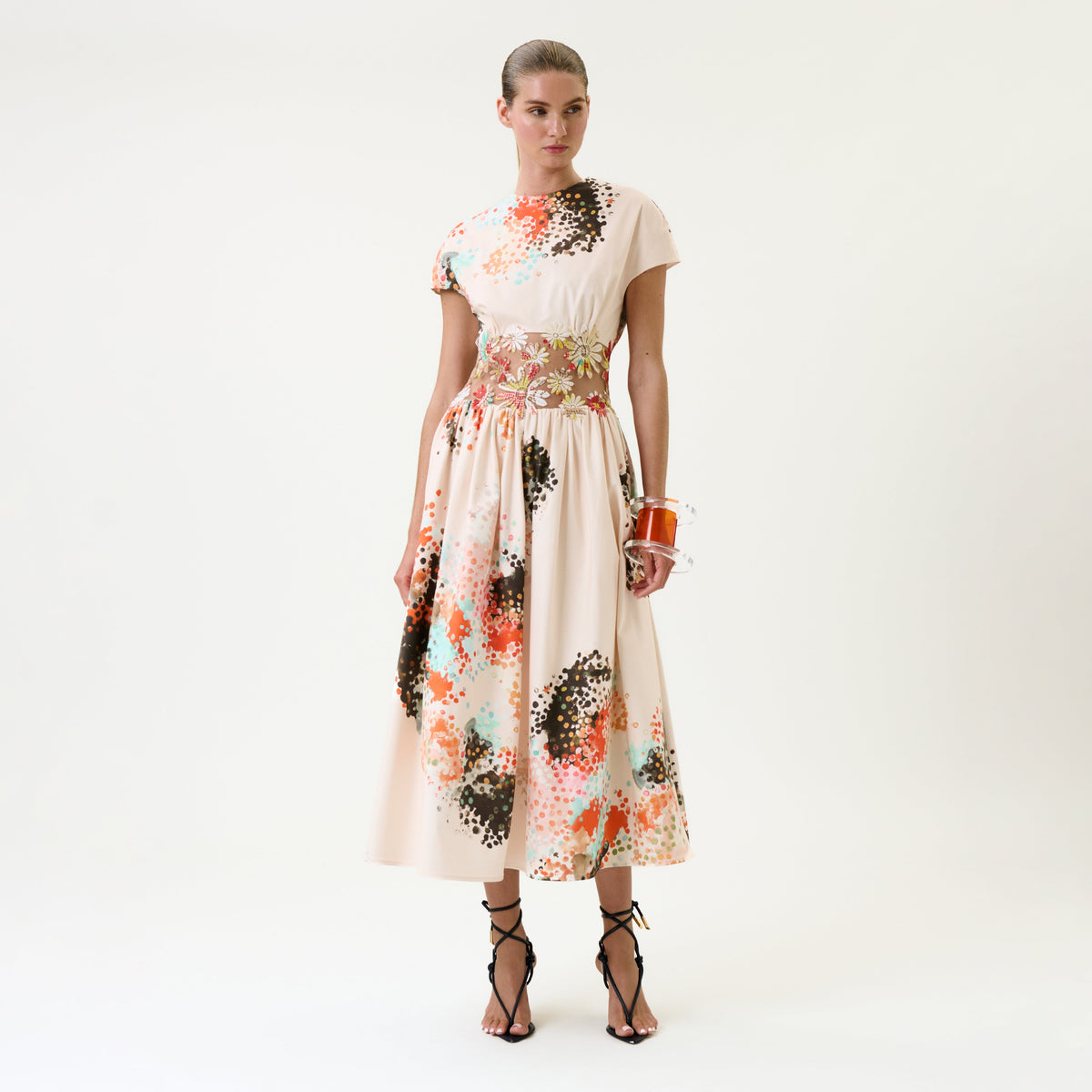 A woman stands against a plain white background wearing the Macaria Dress—a cream-colored, ankle-length dress with colorful abstract patterns—paired with black strappy heels and a small orange bag.