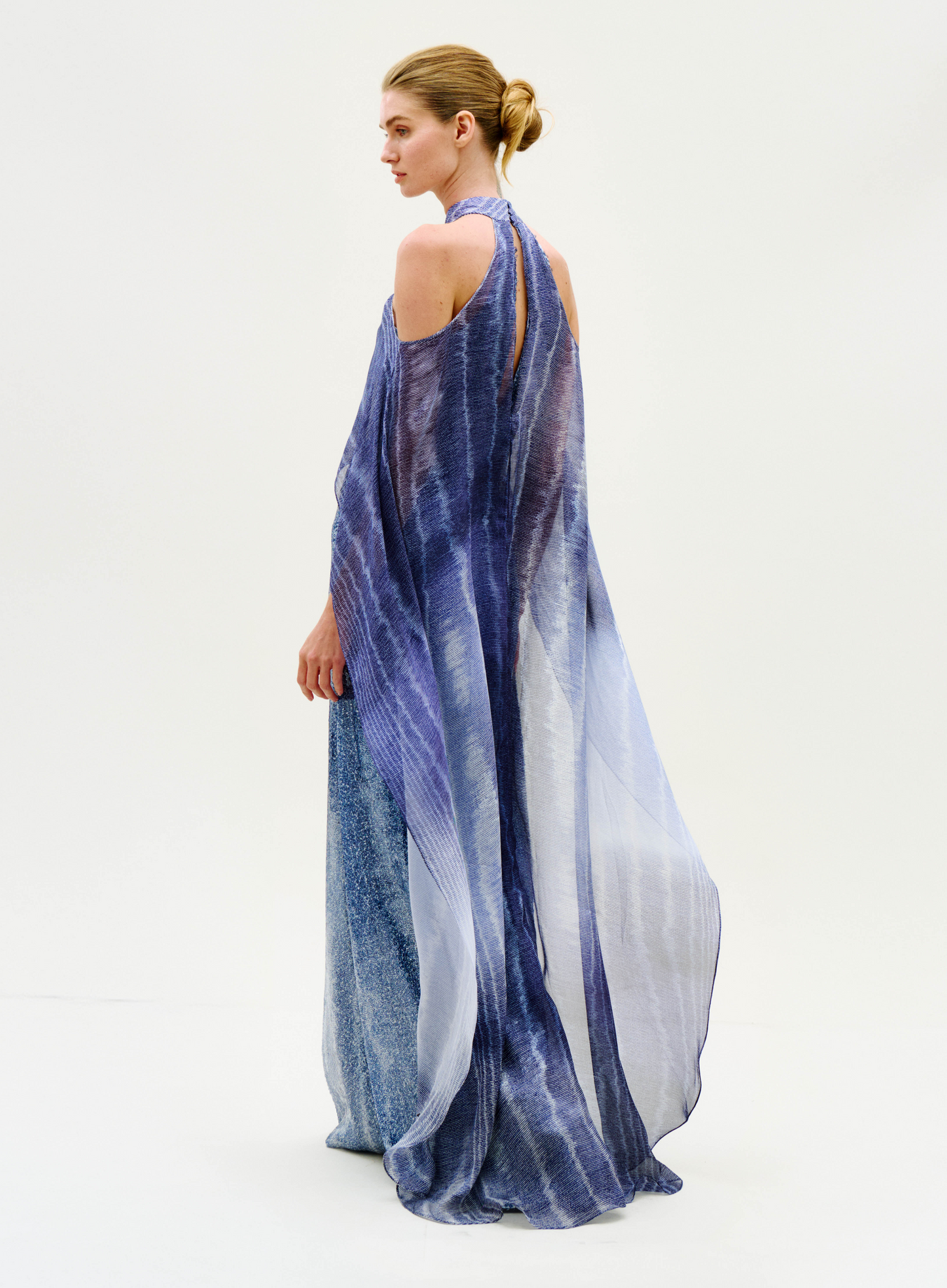 Wearing the Serenela Dress, a person with hair in a low bun stands against a plain, light background, looking to the side. The flowing, sheer dress features open shoulders and comes in a blue-purple hue.